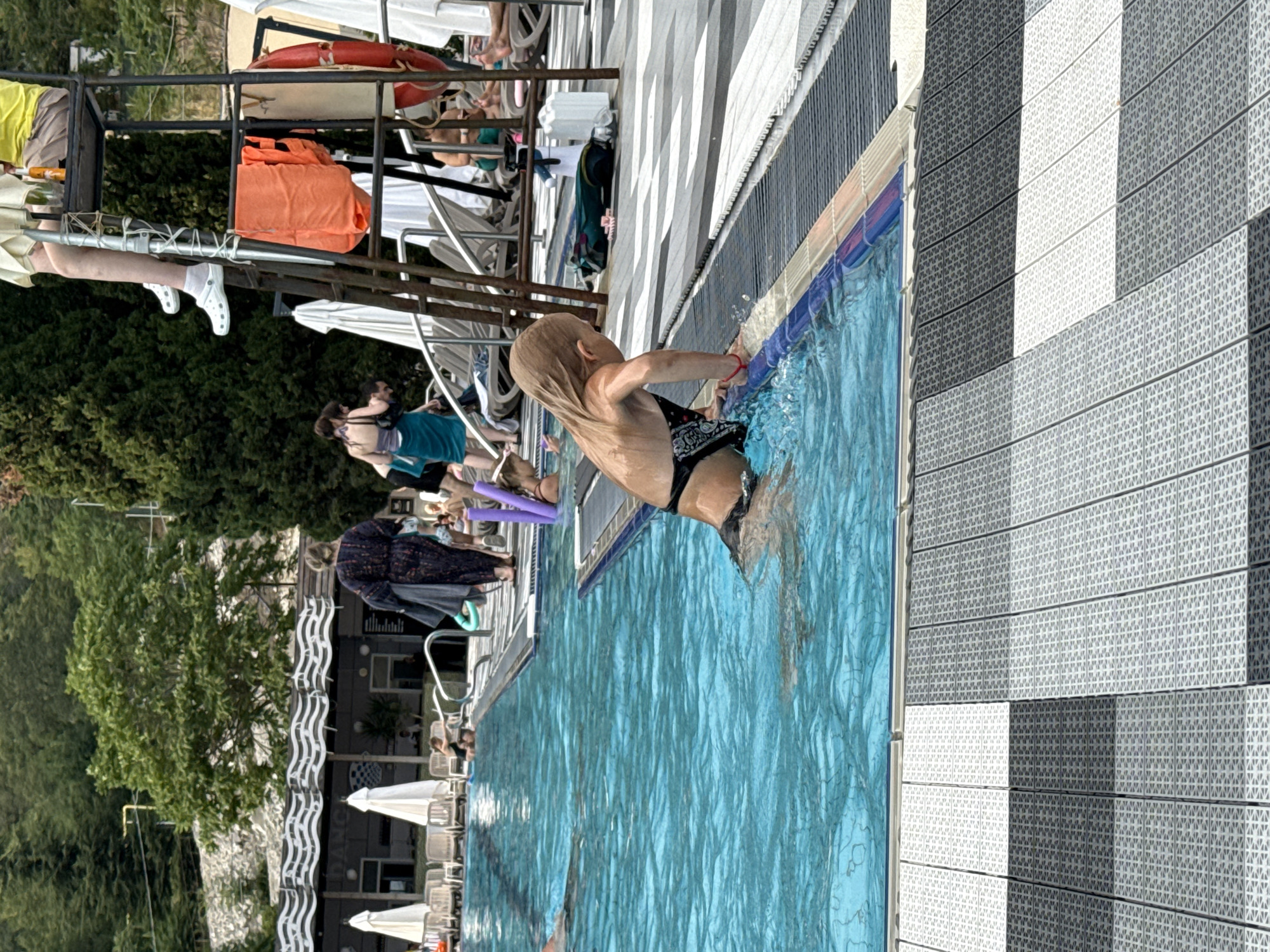 In this horizontal, eye-level shot, a young child with blonde hair is partially submerged in a bright blue swimming pool, holding onto the edge. The child is wearing black swim trunks with a white pattern. The pool's edge is lined with blue and beige tiles. The surrounding area features a tiled deck with a checkered pattern of light and dark gray tiles. In the background, several people are visible, some sitting on lounge chairs under white umbrellas. Trees and a building with windows and a sign are also present. The lighting suggests it is daytime, with sunlight casting shadows. One of the cropped images shows a red life preserver, and another shows a life vest. The overall scene depicts a resort or public swimming area, possibly in Yalta, Ukraine, where people are enjoying a sunny day at the pool.