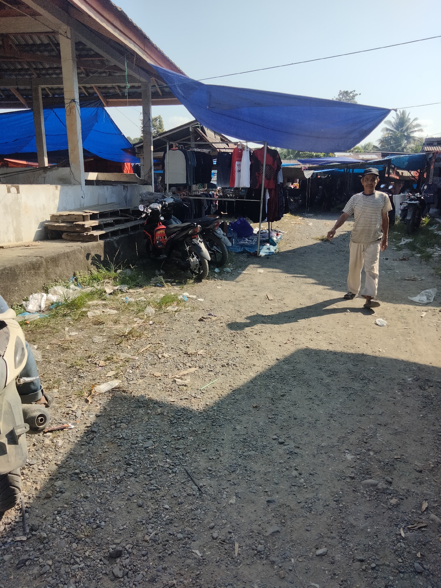 An outdoor market scene is depicted in Indonesia. In the foreground, a dirt and gravel pathway leads into the scene. A person wearing a striped t-shirt and loose pants is walking towards the viewer, casting a long shadow. The market stalls are visible in the background, some covered with blue tarps, displaying clothing on racks. Several motorcycles are parked near the stalls. The environment appears to be sunny with clear skies.