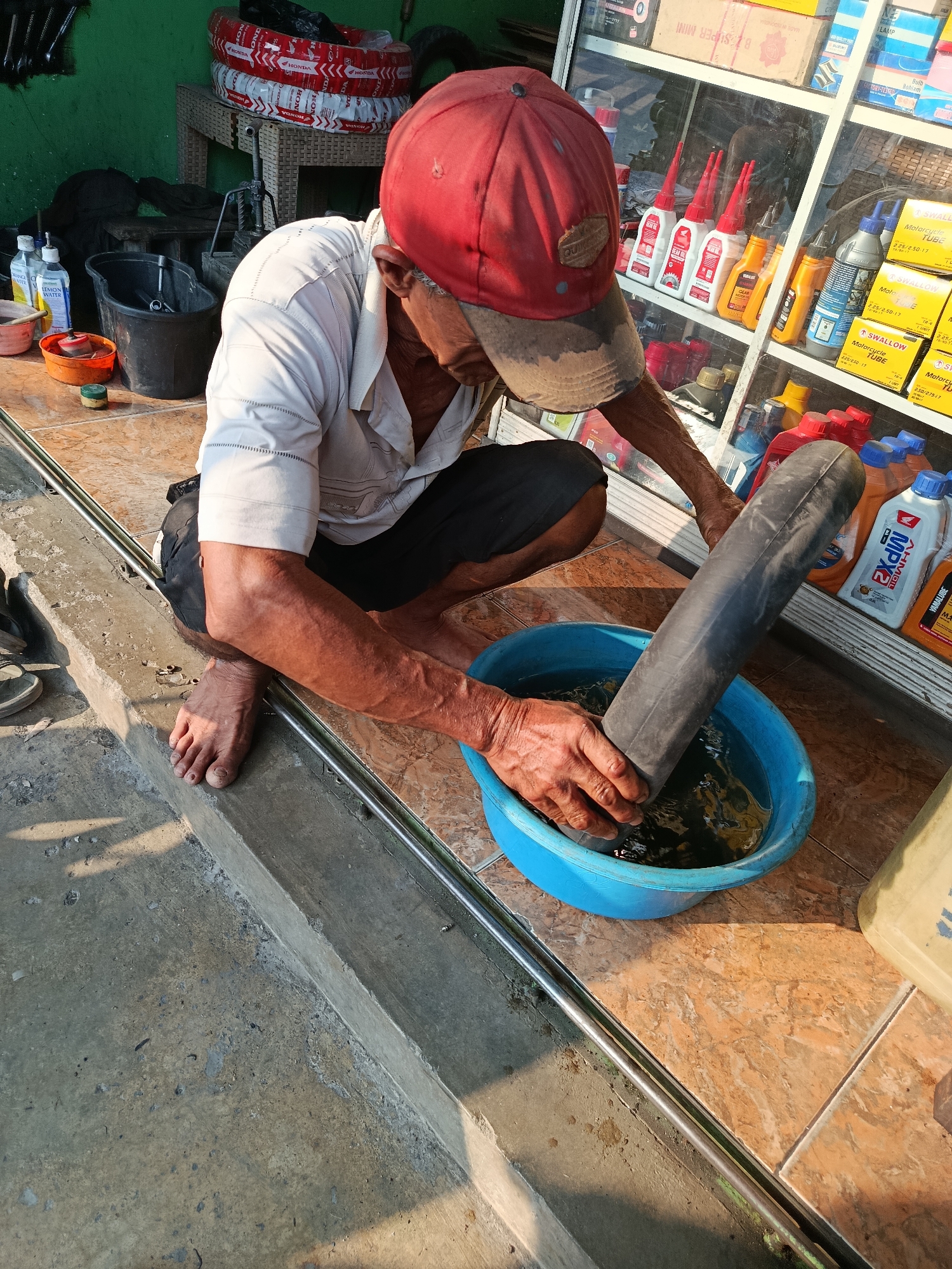 A man wearing a red cap and a light-colored shirt is crouching down, holding a black inner tube that is submerged in a blue tub filled with water. He appears to be checking for leaks in the inner tube by looking for bubbles. The scene is taking place on a tiled floor, with a concrete ledge in the foreground. Behind him, there is a display shelf filled with various bottles and products, including what appear to be engine oils and cleaning supplies. A stack of tires is also visible in the background. The environment suggests a small shop or repair area, possibly a motorcycle repair shop, located outdoors or in a semi-outdoor setting, given the sunlight casting shadows. The presence of products with Indonesian labels, such as "Honda" and "Yamalube," indicates the location is likely in Indonesia. Considering the overall aesthetic and common types of shops found in Palembang, Indonesia, this scene is consistent with a roadside repair shop or a small retail establishment selling automotive parts in a local market area of the city.