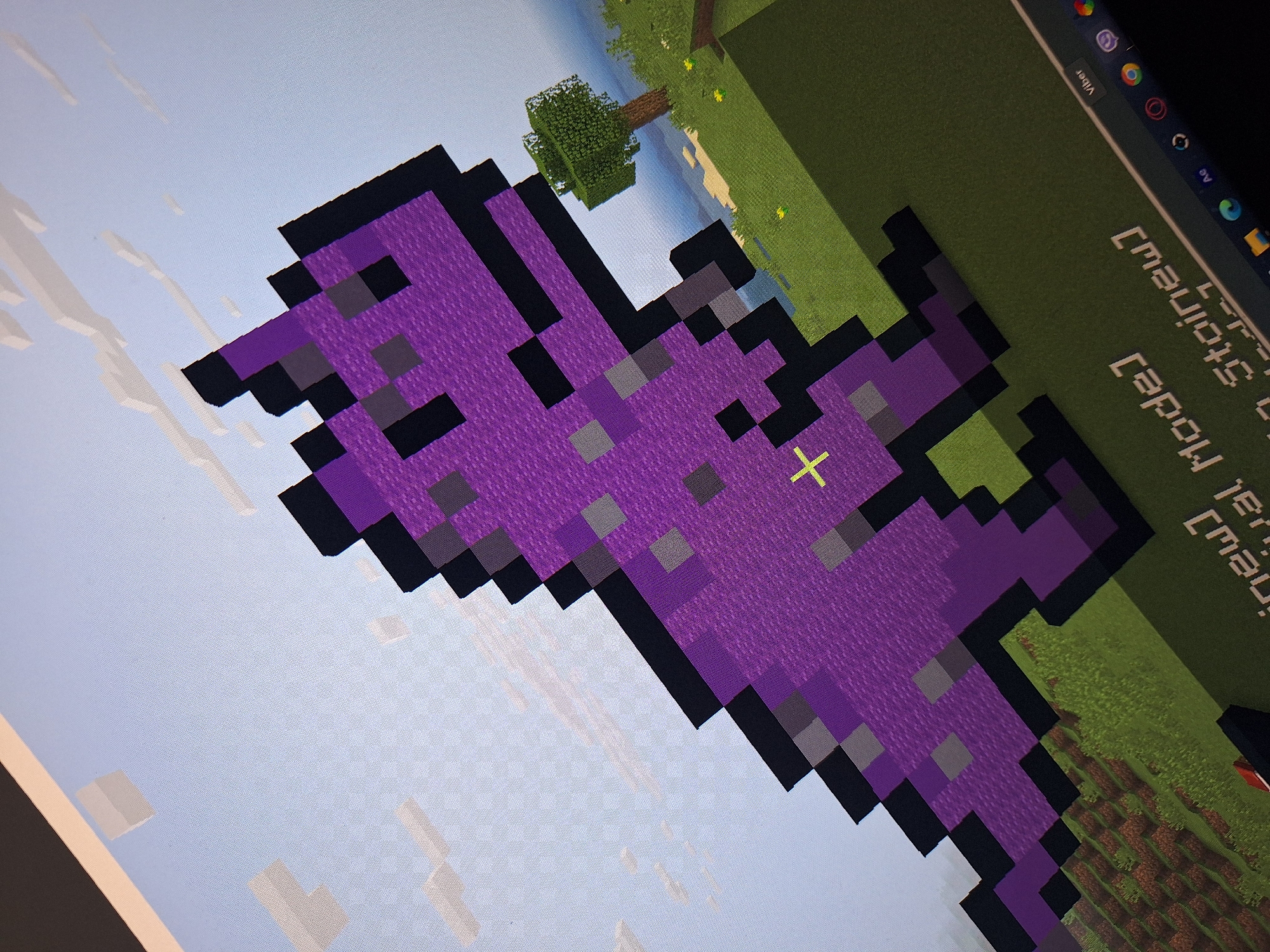 The image shows a computer screen displaying a pixelated purple dinosaur in a Minecraft game environment. The dinosaur is a prominent subject, rendered in shades of purple and gray with black outlines. The game world features green grassy terrain, brown dirt patches, a sandy area, a tree, and a light blue sky with white pixelated clouds. In the lower right corner of the screen, a portion of a computer taskbar is visible, displaying icons for applications like Viber, Chrome, and Adobe. The image itself is a photograph of the screen, taken from an angle, suggesting it is being viewed on a physical monitor. There is no indication of the city of Varna, Bulgaria, or any other specific geographical location.