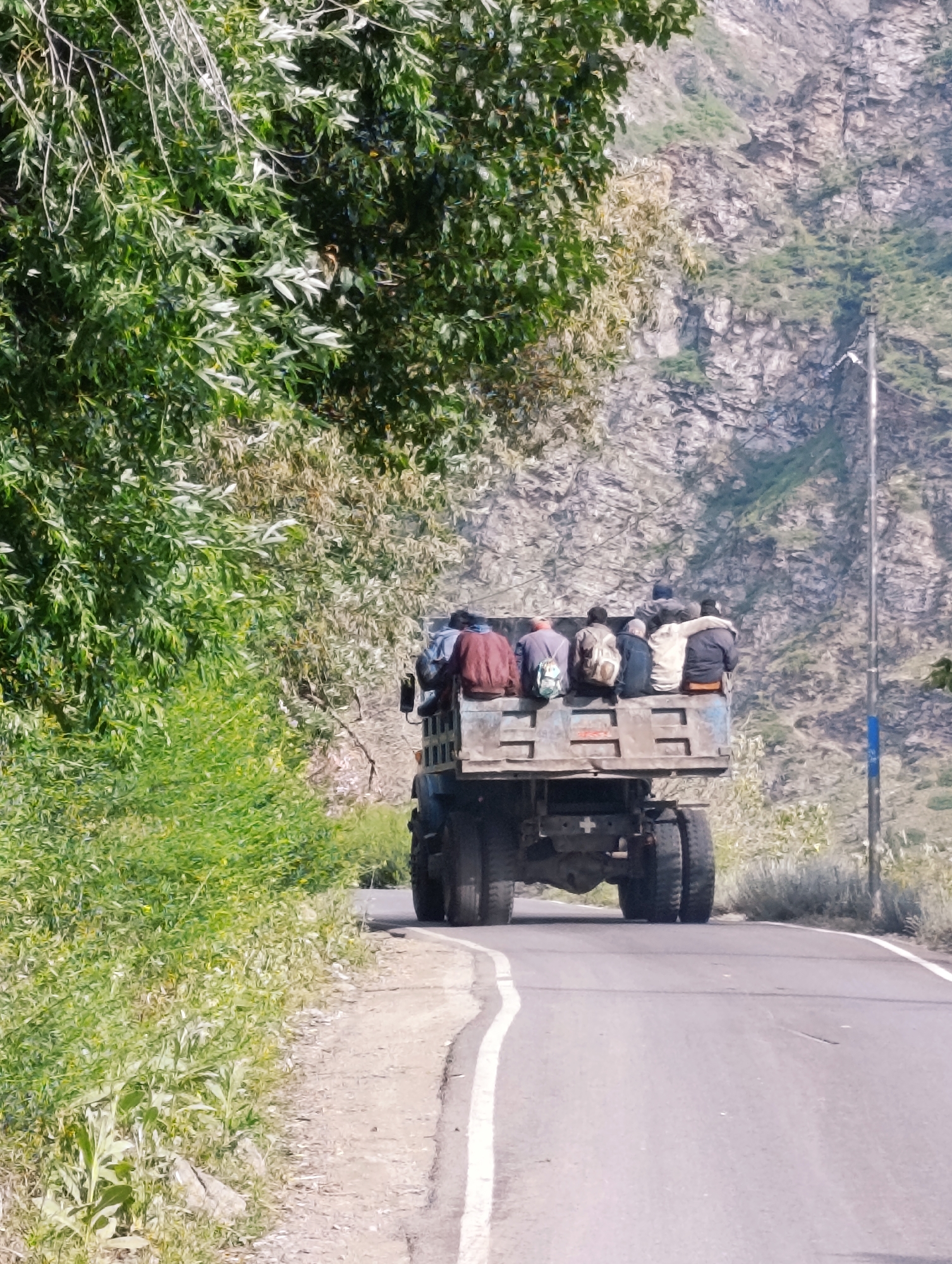 In Kirting, India, a flatbed truck is captured in motion on a paved road that winds through a mountainous, vegetated terrain. The truck's bed is filled with eight individuals, appearing to be passengers, seated on the truck's edge. They are dressed in casual clothing. The truck is traveling away from the viewer, and its rear wheels are visible. The environment includes lush green trees and foliage on the left side of the road, and a rocky, mountainous hillside on the right. A utility pole with a blue marker is situated on the right side of the road, near the truck. The lighting suggests it is daytime.