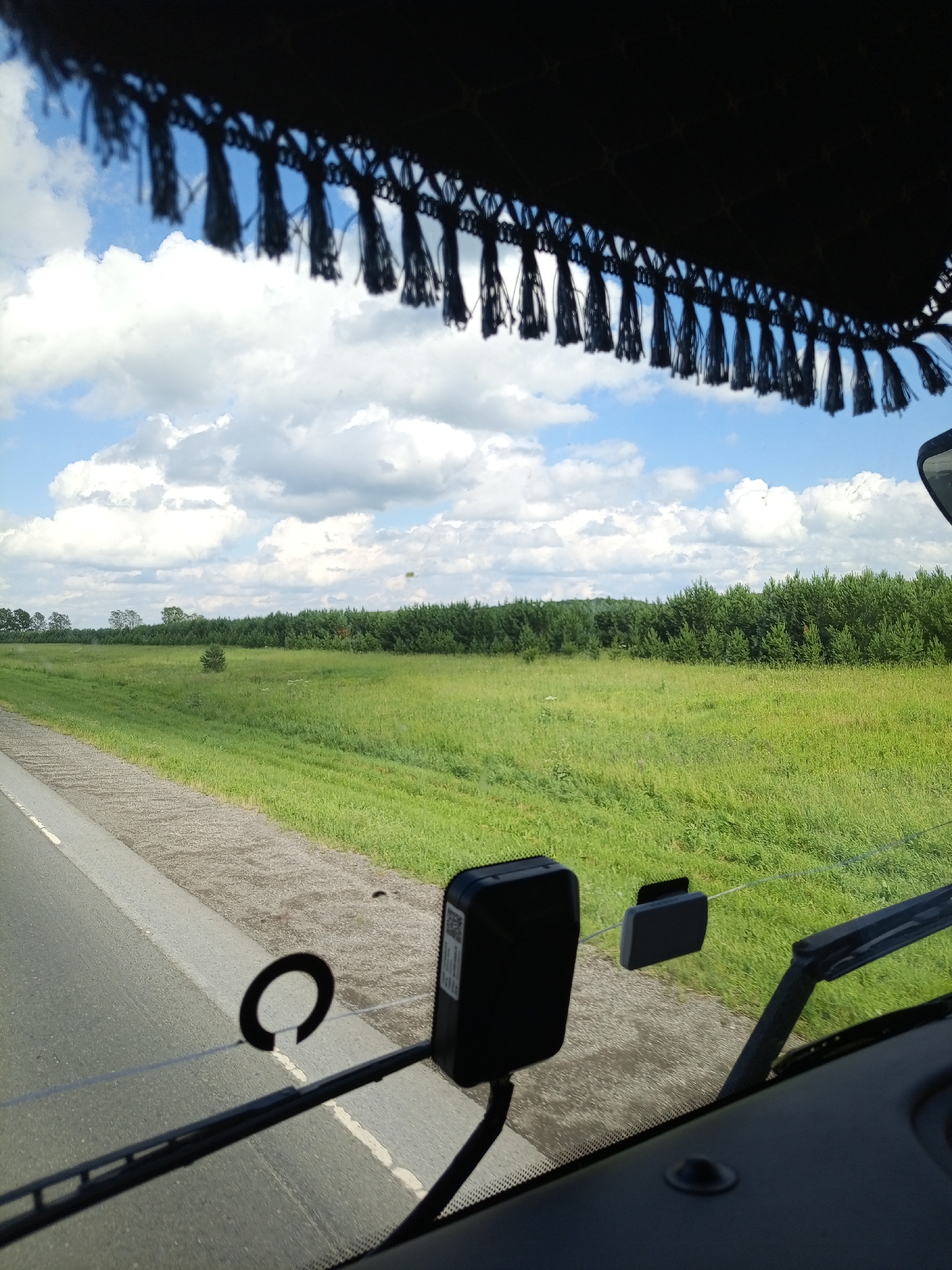 The image, taken from the interior of a vehicle, shows a view through the windshield on a bright, cloudy day. The foreground is dominated by the dashboard and windshield wipers, with a black tassel fringe visible at the top of the windshield, suggesting the vehicle is likely a truck or RV. A circular object, possibly a sticker or suction cup, is affixed to the windshield. To the left, a dark rectangular device with a QR code is mounted.
Beyond the windshield, a vast, green meadow stretches towards a dense line of dark green trees that form the horizon. The meadow appears lush with grass and some wildflowers. In the distance, a small, solitary tree stands in the middle of the field. The sky is a vibrant blue, dotted with large, fluffy white clouds. A small, light-colored object, possibly a kite or a drone, is visible high in the sky above the meadow. The road is visible on the left side of the image, with a paved shoulder. The overall impression is a serene, rural landscape.
The provided metadata indicates the image was taken in Verkh-Chebula, Russia. This context suggests the scene is likely a rural road in the Tomsk Oblast region of Russia.
