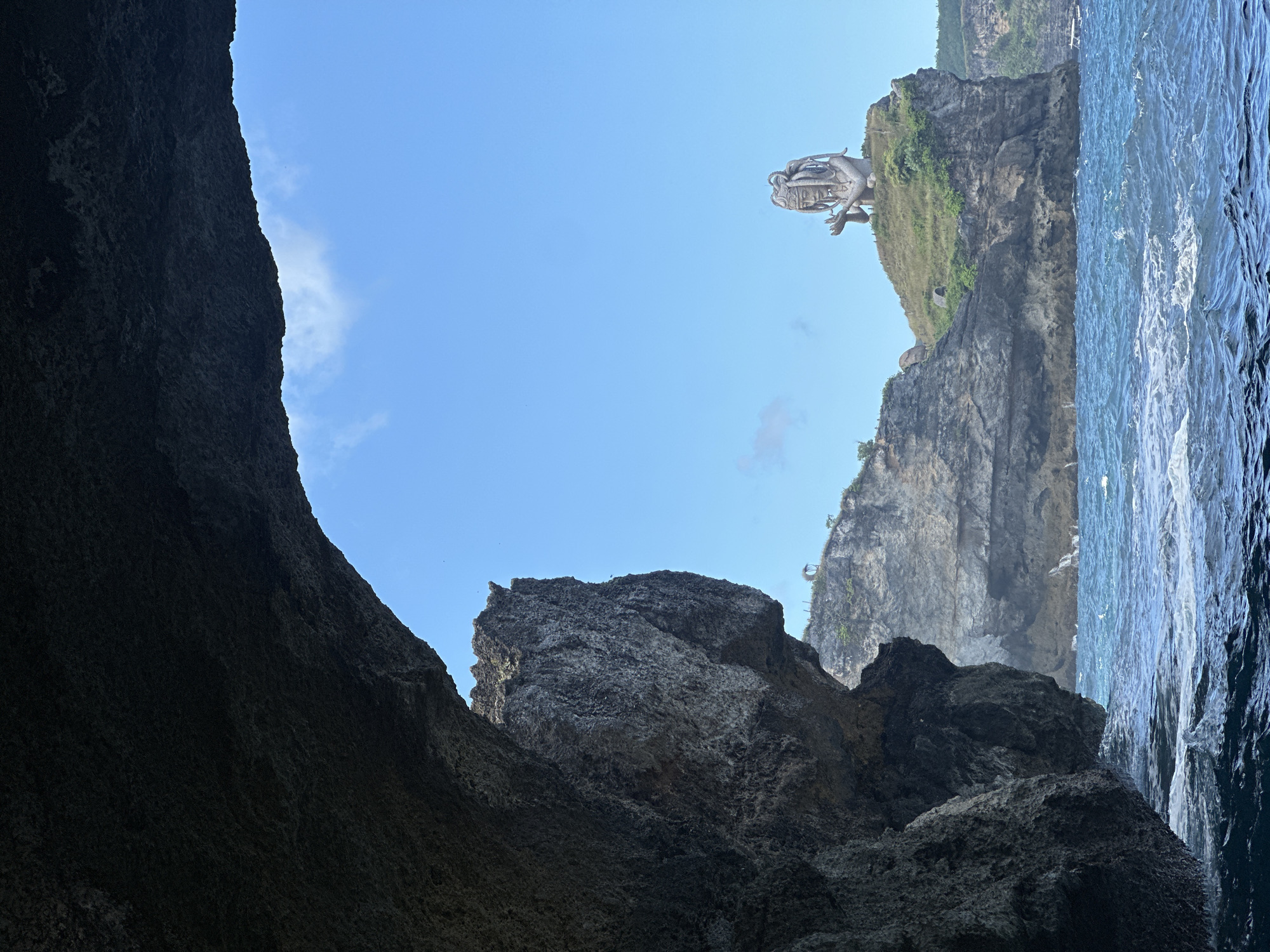 The image shows a view from inside a dark cave looking out toward a rocky cliffside and the ocean. On the cliff, there is a statue of a dragon-like figure. The sky is blue and mostly clear. The location is Nusa Penida, Indonesia.