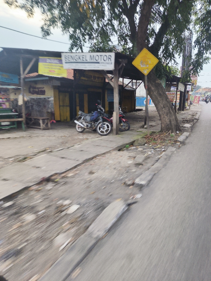 The image shows a street in Cileunyi, Indonesia. It's a busy street with a lot of traffic. The street is lined with shops on both sides. On the left side of the image, there's a shop called "Sengel Motor" and another shop behind it. A sign with a yellow diamond with a symbol on it hangs from a tree. The tree is partially obscured in the image. The street is paved with concrete and has a sidewalk on the left side, which is covered in dirt and debris. There are two motorcycles parked on the right side of the image. The road appears to be busy with cars and motorcycles. The image was likely taken on a sunny day, as there is a lot of light.