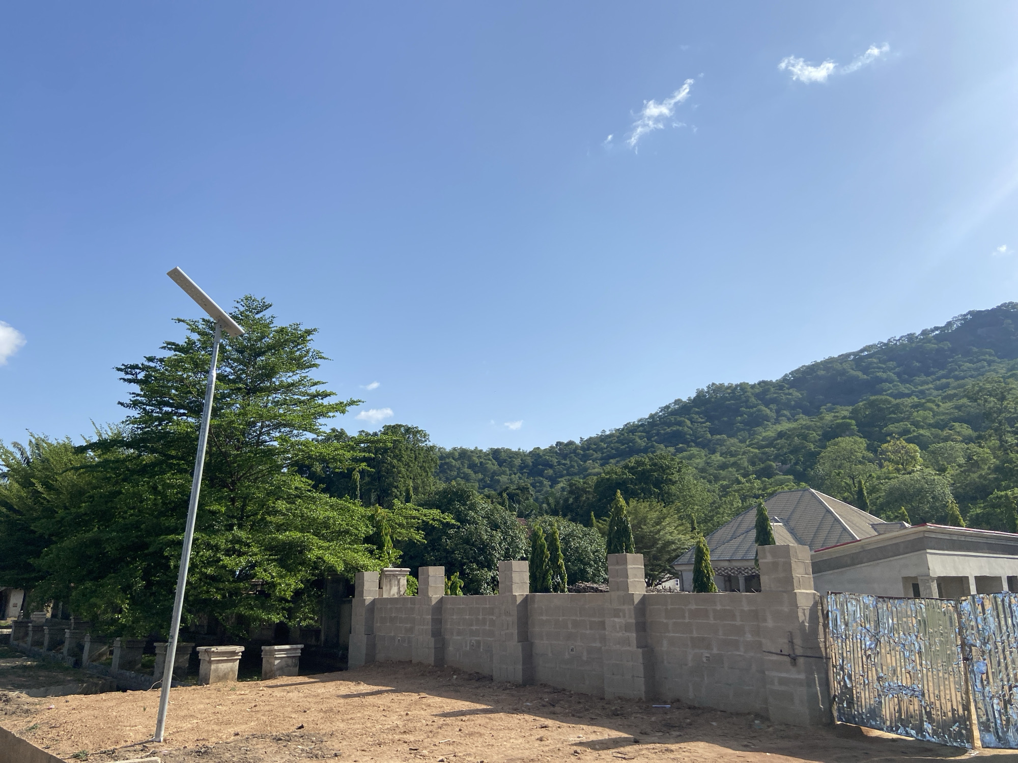 The image depicts a construction site in what is likely a developing area of Mararaba, Nigeria. The foreground shows a dusty, dirt ground leading up to an unfinished cinder block wall. The wall runs horizontally across the middle of the frame. Behind the wall, a partly-constructed house with a visible roof is seen among lush green trees. A steep, densely-forested mountain rises in the background. A modern solar-powered street light stands to the left, partially obscured by a tall tree. The sky is clear and bright blue with scattered clouds. The overall impression is that of construction activity in a location where urban development meets a natural, hilly landscape. The partially constructed wall and gate along with building suggest a gated residential or commercial development. No people are visible in the scene.