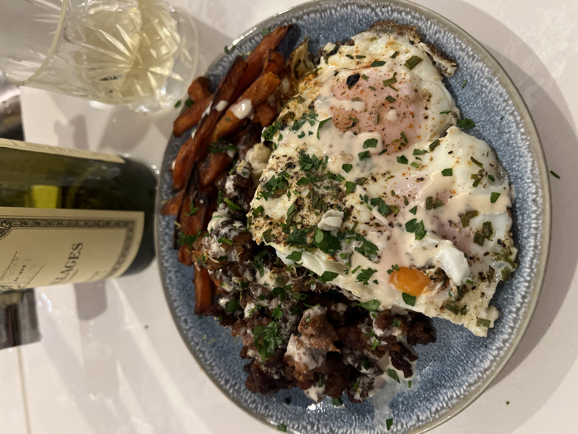 The image depicts a plated meal on a light blue plate. The meal consists of sweet potato fries with white drizzle, a dark, ground meat mixture with white drizzle and parsley, and two fried eggs with parsley on top. A partially visible wine bottle and a glass of white wine are positioned next to the plate. The background is a white surface. The location is unknown and cannot be confirmed as London, United Kingdom based on the visual information provided.