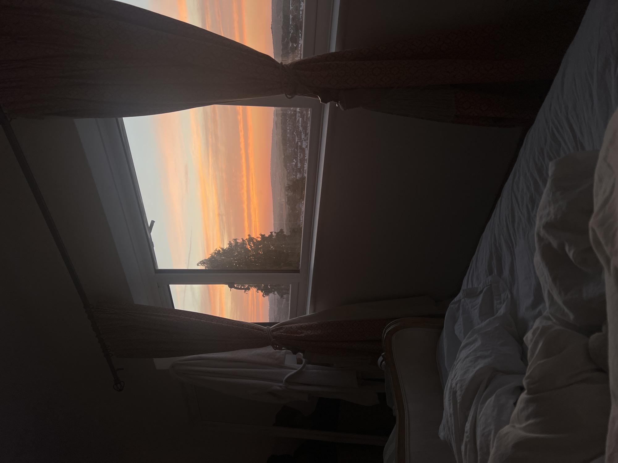 A bedroom interior overlooks a sunset in Brighton, United Kingdom. The foreground features a rumpled bed with white linens. To the left, a white bathrobe hangs on a door or wall. The window, framed by patterned curtains, reveals a panoramic view of the city bathed in the warm hues of the setting sun. The sky is a vibrant gradient of orange, pink, and pale blue. Silhouetted against the sky are trees and the distant cityscape.