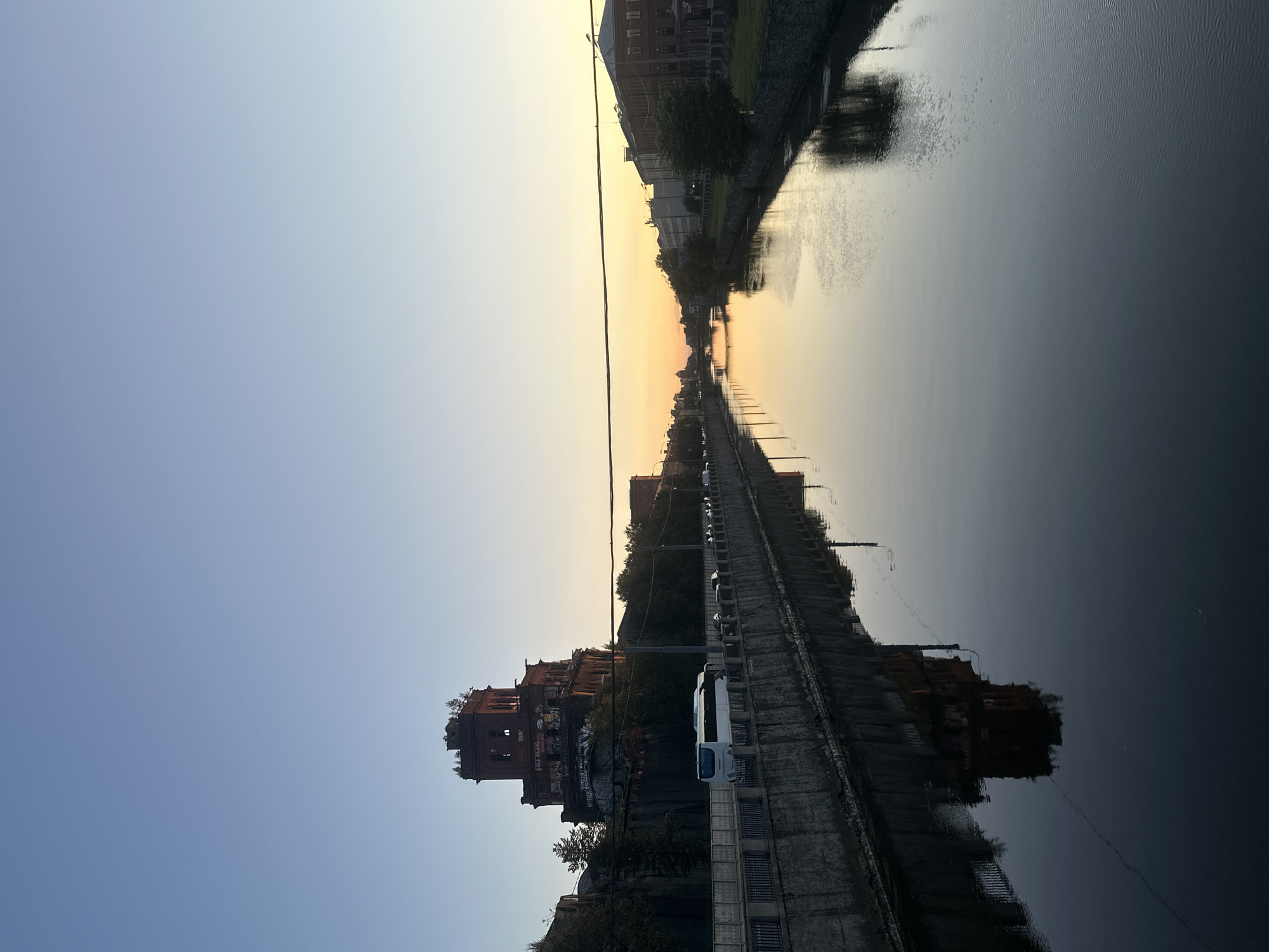 The image captures a tranquil canal scene in Saint Petersburg, Russia, during what appears to be sunset or early dawn. The sky transitions from a clear, pale blue at the top to a warm, soft yellow and orange hue near the horizon, indicating the time of day. A prominent, derelict brick tower, possibly an old industrial building, stands on the left bank of the canal, adorned with graffiti. Adjacent to it, a white bus and several cars are visible on the road lining the canal. The right bank features a more typical urban landscape with buildings and trees. The still water of the canal reflects the sky, the buildings, and the imposing tower, creating a symmetrical and serene composition. A thin, dark horizontal line, likely a cable or wire, stretches across the upper part of the frame. The canal walls are constructed of stone.