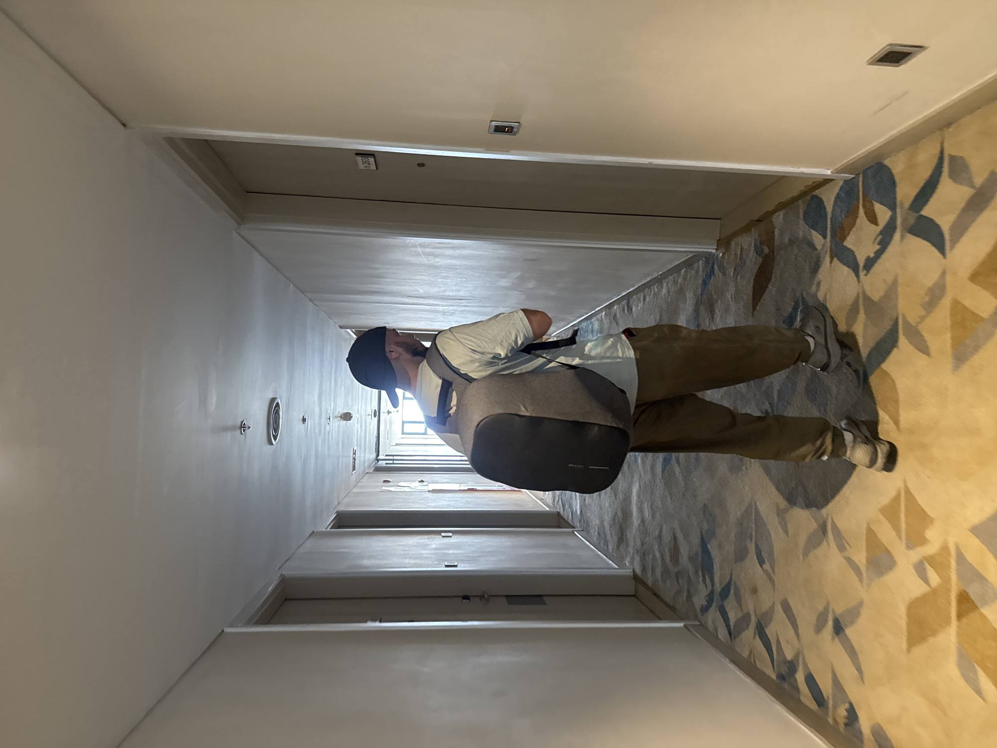 A photograph shows a person standing in a hotel hallway. The hallway walls are cream-colored, while the carpet has a design with blue and beige hues. The person wears a black cap, a white shirt, and khaki pants, carrying a gray backpack. The lighting fixtures and door number plaques on the wall are visible. The specific location in Đà Nẵng, Vietnam, is not determinable from the image.