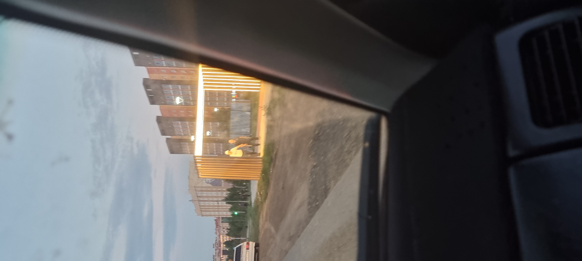 An eye-level shot, taken from inside a vehicle, captures a street scene in Aktobe, Kazakhstan. In the foreground, the dashboard and windshield frame the view. To the left, a white station wagon is parked on a dirt road. Beyond it, a row of trees and a green traffic light are visible. In the midground, two men are walking towards a modern, illuminated archway. The archway is constructed with vertical wooden slats and illuminated with bright lights, creating a striking visual contrast against the dusk sky. Behind the archway, a multi-story building with a patterned facade stands prominently, with other buildings of similar architecture extending into the background. The sky above is a pale blue with scattered clouds, indicating either early morning or late evening. The overall atmosphere is calm and suburban, with a hint of urban development.