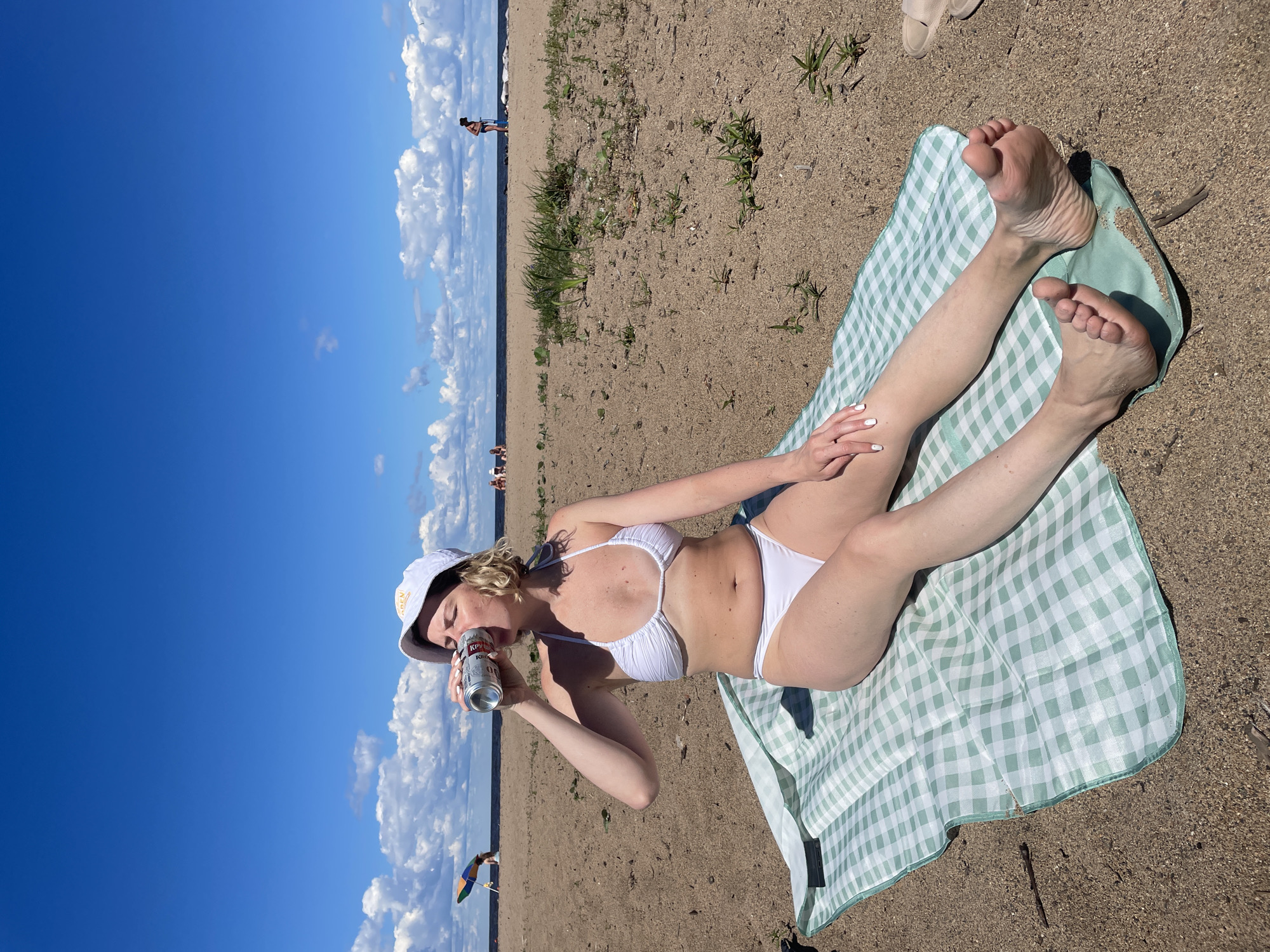 An overhead medium shot shows a woman sitting on a beach blanket on a sandy beach. The woman is wearing a white bikini and a white bucket hat. She is holding a can and drinking from it with her right hand. Her legs are extended out in front of her. The blanket is green and white checkered. The background shows a beach with a few people in the distance, a body of water, and a blue sky with white clouds. The scene appears to be taking place on a sunny day. There is no identifiable landmark linking this image to the city of Belozersk, Russia.