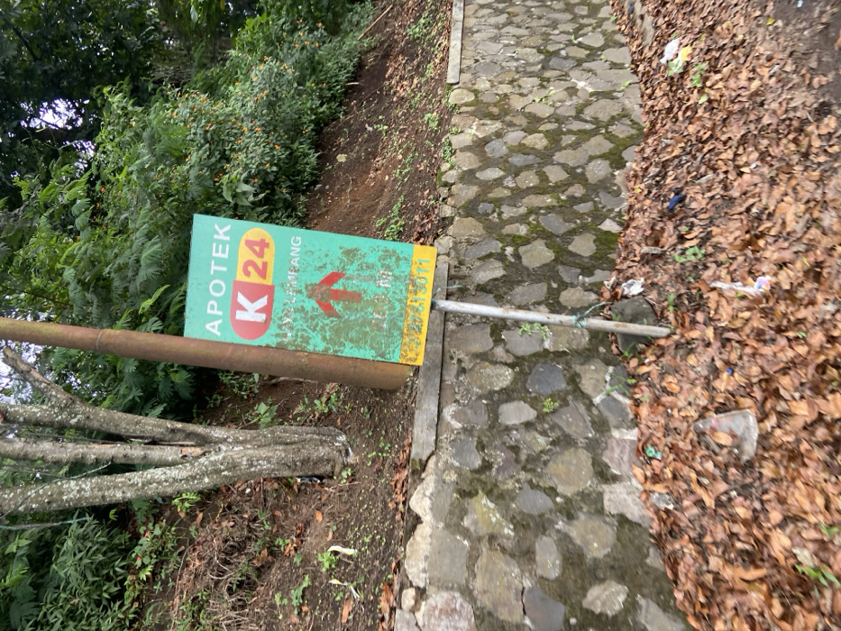 A green and yellow sign with white lettering is mounted on a metal pole, attached to a concrete post. The sign reads "Apotek K24" with an arrow pointing upwards. The sign is partially obscured by a tree trunk. The sign is located on a dirt slope with a stone wall. There are brown leaves on the ground. The location is on a street in Lembang, Indonesia, at the coordinates -6.828, 107.607. The sign is likely for a local pharmacy.