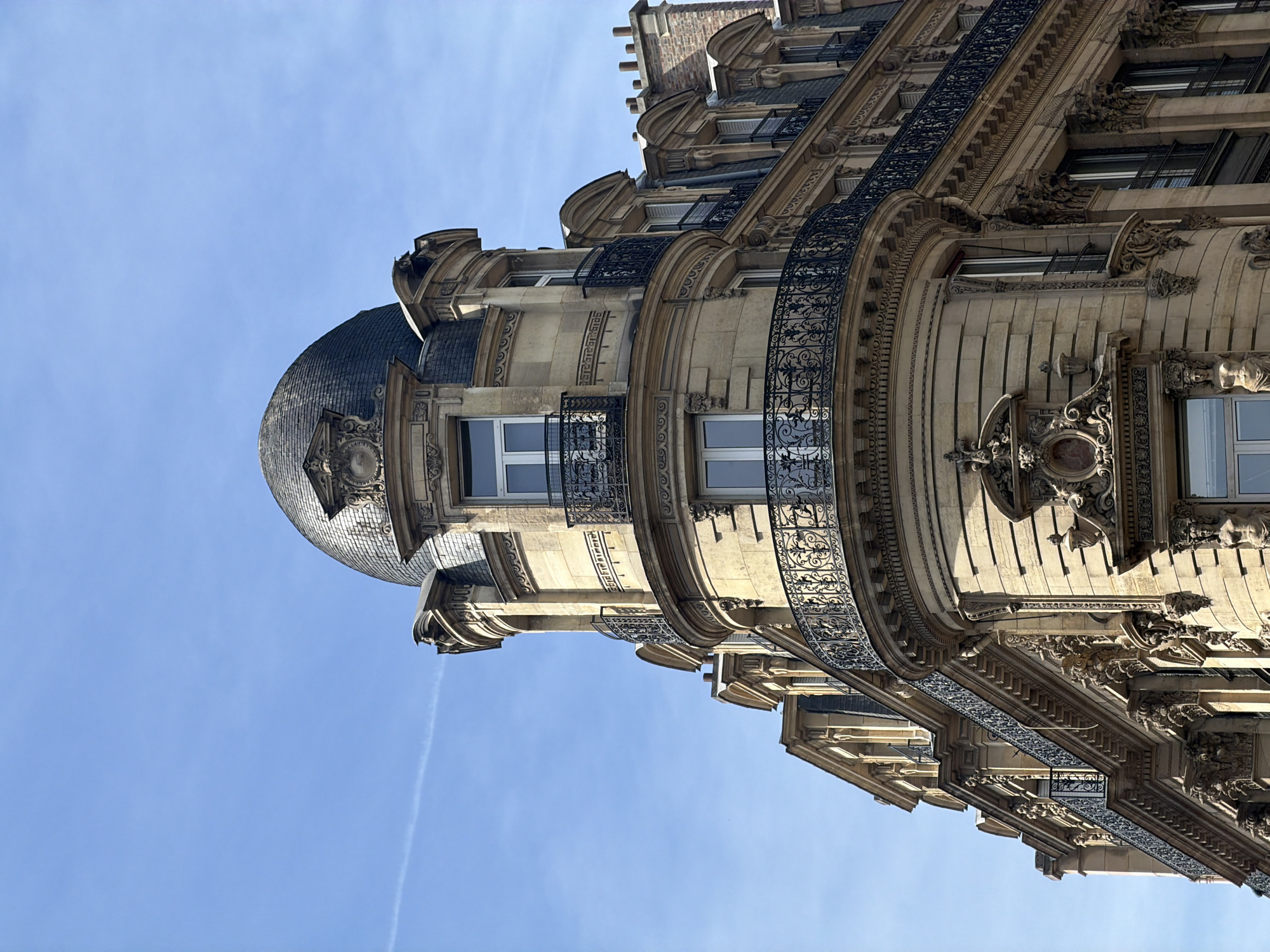 The image presents a striking low-angle view of an ornate, multi-story building exterior set against a clear, bright blue sky, typical of Parisian architecture. The building features a prominent rounded corner that culminates in a dark, possibly slate-tiled dome or cupola at its uppermost visible point.
The facade is richly detailed with intricate stonework, showcasing classical elements such as decorative moldings, cornices, pilasters, and various carved embellishments like garlands, rosettes, and other sculptural motifs. Several floors are visible, each with windows, some featuring white frames, and balconies. These balconies are adorned with elaborate black wrought-iron railings, characterized by intricate scrollwork and decorative patterns, which stand out against the light-colored stone.
The bright daylight illuminates the building, casting crisp shadows that emphasize the architectural depth and texture. A faint white contrail is visible across the otherwise clear blue sky, indicating good weather conditions, likely morning or afternoon. The grandeur, detailed ironwork, and distinctive stone facade are hallmarks of 19th-century Haussmannian architecture, firmly placing the scene in Paris, France. No people or activities are visible, and there is no discernible text. The image captures the timeless elegance and architectural splendor of the city.