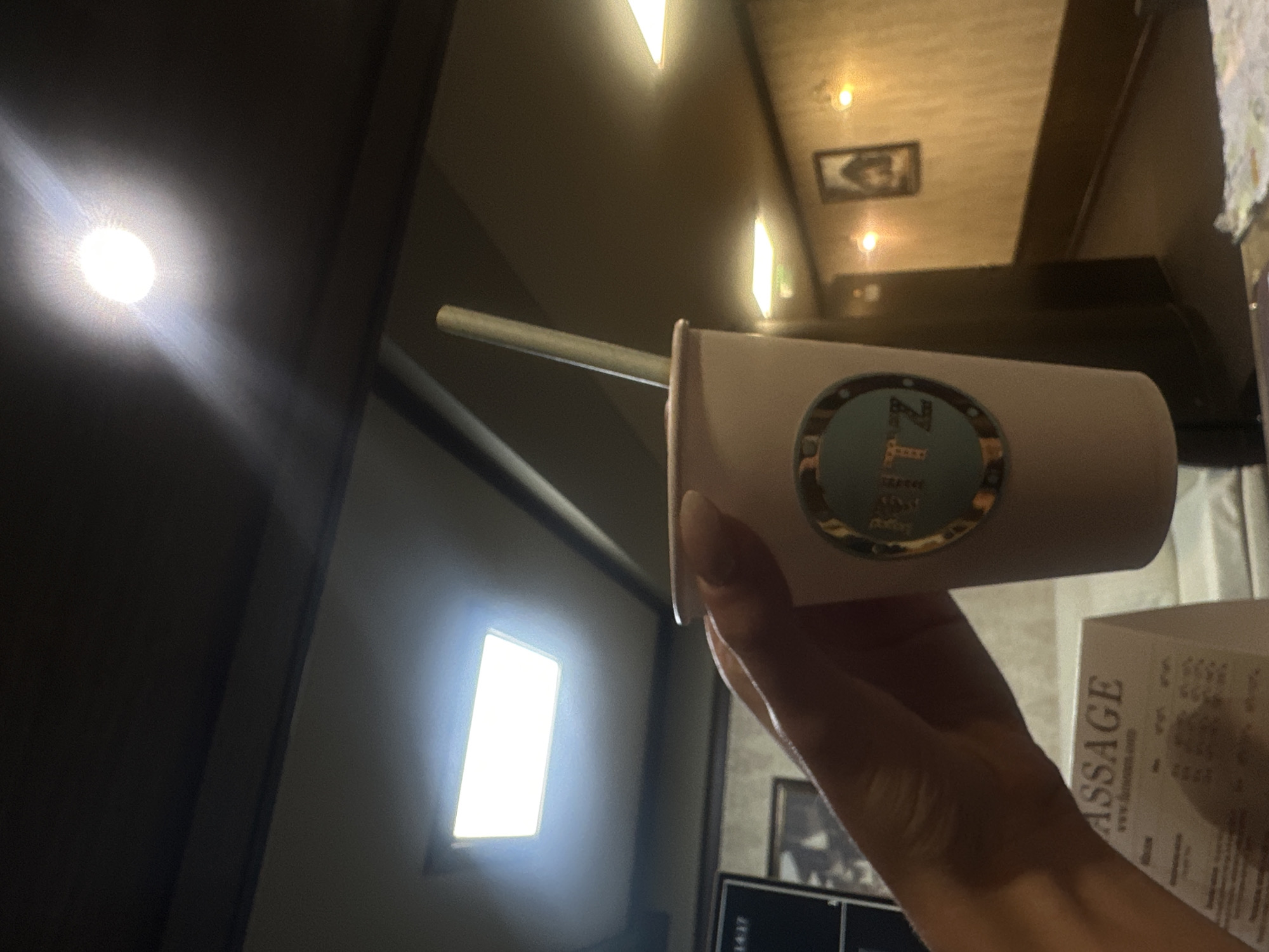The image displays a close-up of a right hand holding a light pink disposable cup with a light-colored straw. The cup features a circular teal and gold logo with the letters "TZ" prominent, suggesting "Relax TZ Massage & Spa Center." The hand exhibits long, light-colored manicured fingernails.
The scene is an interior shot of the Relax TZ Massage & Spa Center, located at ul. "Lachezar Stanchev" 5, 1756 zh.k. Darvenitsa, Sofia, Bulgaria. This identification is supported by the partial visibility of a white tent card in the lower-left foreground, which prominently displays "MASSAGE" and "www.relaxam.com." The tent card rests on a light-colored, possibly white, upholstered surface.
The background is dimly lit. The ceiling is dark, featuring a bright rectangular LED panel light on the left and a circular recessed light source towards the center-right, emitting strong illumination. To the right, a hallway or corridor recedes into the distance. Its walls are a light beige with a textured appearance, accented by a dark wainscoting or baseboard. Several framed pictures are hung along this wall, individually illuminated by small, warm-toned sconce lights. A dark, rectangular object, possibly a door or cabinet, is partially visible on the left side of the hallway.