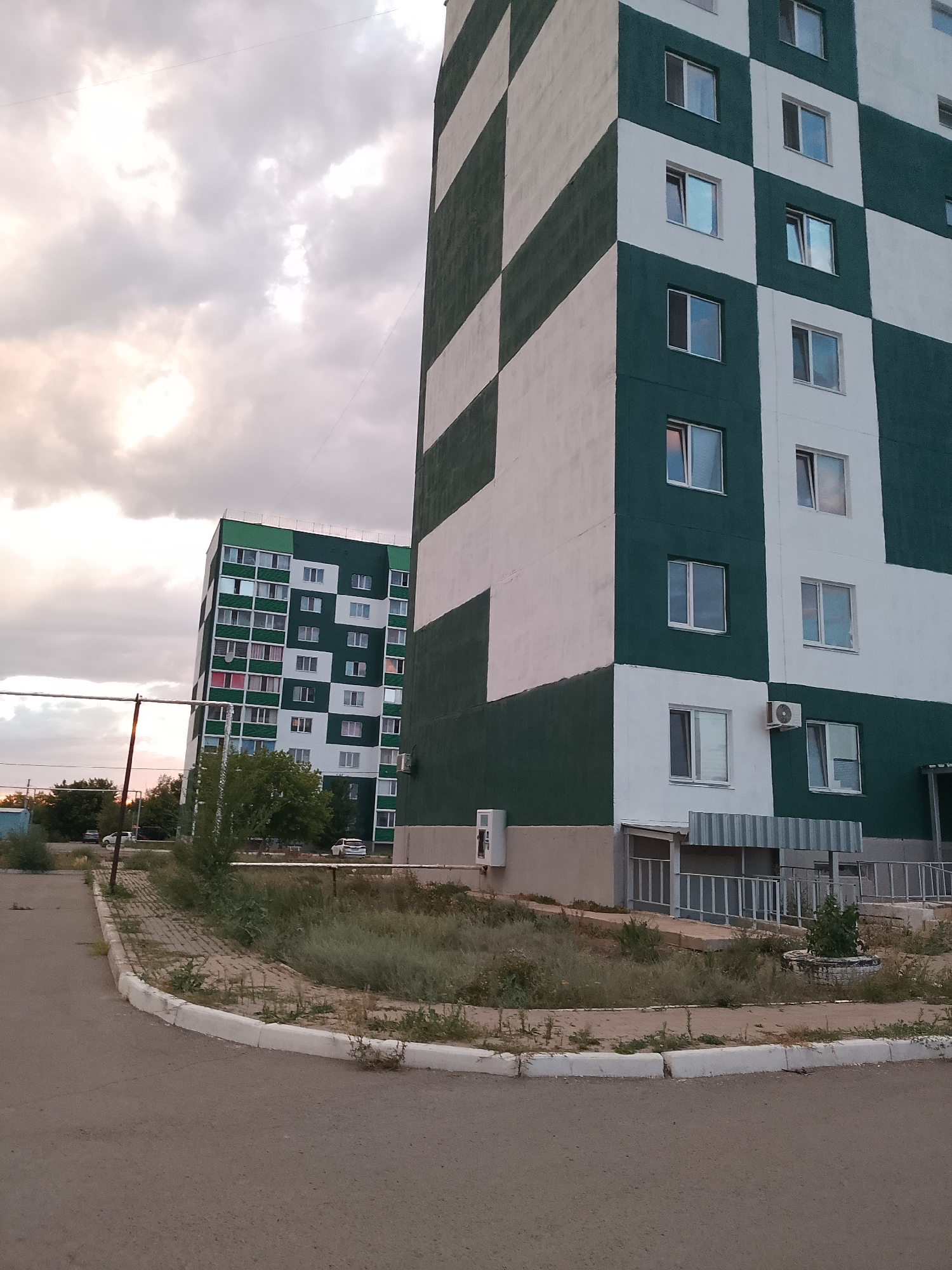 The image depicts an outdoor scene in the city of Abay, Kazakhstan, focusing on two residential apartment buildings. The building in the foreground is tall and features a distinctive checkerboard pattern of dark green and white squares. Numerous windows are visible on its facade. To the left, another apartment building is visible, also with a green and white color scheme, though with a different pattern. This building appears to be slightly tilted. The surrounding environment includes a paved road or pathway in the lower part of the image, with a curved concrete curb and overgrown dry grass and weeds. A metal pipe runs horizontally across the mid-left portion of the frame, supported by poles. Trees are present in the background, and several cars are parked near the buildings. The sky above is cloudy, with muted light suggesting it is either early morning or late evening. No people are visible in the frame. The scene appears to be a residential area in a post-Soviet urban landscape.