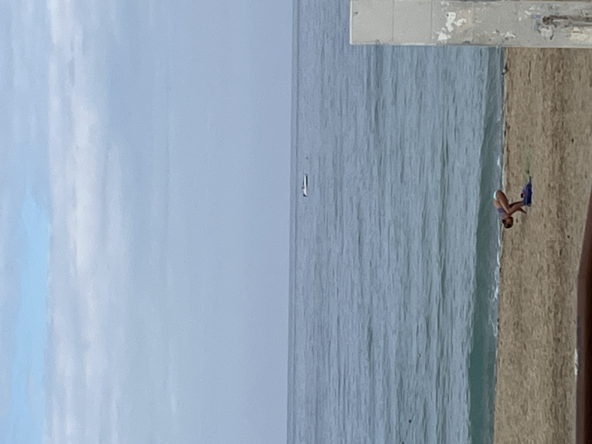 The image shows a scene at a beach in Alanya, Turkey. The environment consists of a wide expanse of pale blue sky, the sea, and a sandy beach. A distant sailboat is visible on the horizon. A woman is on the beach near the water's edge, appearing to be putting on fins. The top portion of a concrete wall or structure is visible in the upper right corner of the frame. A portion of a person's arm is also visible on the right.