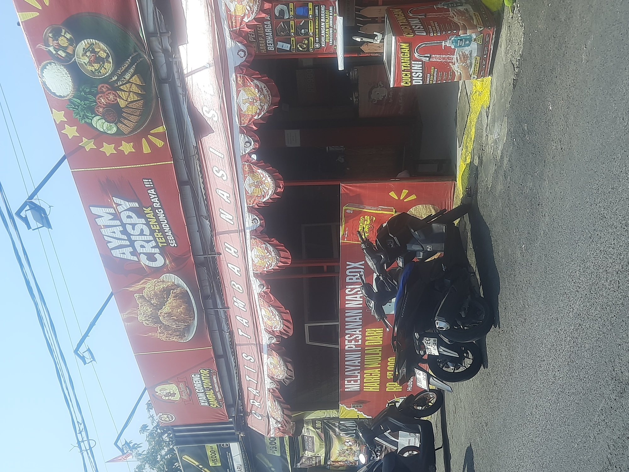This image depicts a storefront in Bandung City, Indonesia. The facade of the building features a large red banner advertising "AYAM CRISPY TER-ENAK SEBANDUNG RAYA!!!" which translates to "The Most Delicious Crispy Chicken in Bandung Raya!!!". Below this, a smaller banner reads "GRATIS TAMBAH NASI SIAP". The storefront appears to be a food establishment, likely a restaurant or food stall, with awnings and signage indicating various food offerings and promotions.
In front of the establishment, several motorcycles are parked on the asphalt road. The area seems to be a busy street, as suggested by the presence of multiple vehicles and the urban setting. Yellow paint marks a line on the road in front of the building. There are also several red banners on the side, one of which advertises "MELAYANI PESANAN NASI BOX HARGA MULAI DARI RP. 17.000" (Serving Nasi Box Orders, Prices Start From Rp. 17,000). On the right side of the image, a cardboard dispenser for handwashing supplies is visible, with text in Indonesian related to handwashing.
The overall scene suggests a commercial area with a focus on food services. The lighting indicates it is daytime, and the sky is visible in the upper portion of the image. The architecture and signage are consistent with typical Indonesian street-level businesses.