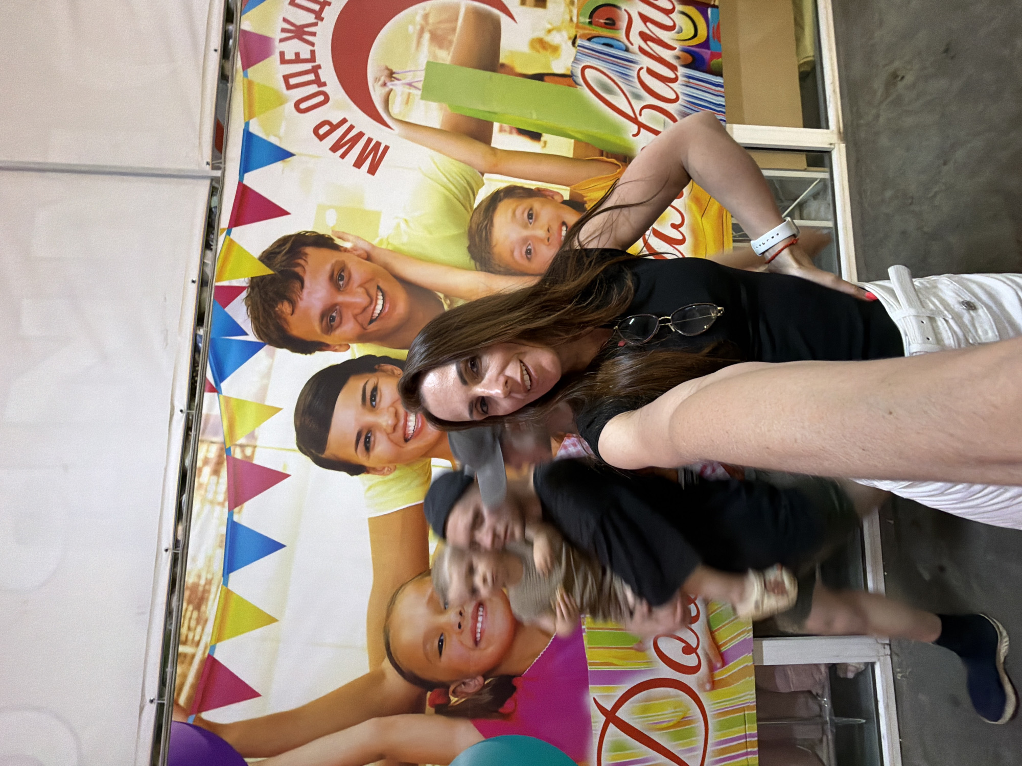 The image is a selfie taken in front of a large banner that appears to be an advertisement. The banner features a family of people, including children, smiling and posing. Text in Russian is visible on the banner, with phrases like "МИР ОДЕЖДЫ" (World of Clothes) and "Бата..." which could be part of a store name or slogan. In the foreground, a woman is taking the selfie, and to her left, a man is holding a young child. Another child and a blurred figure are also visible. The environment seems to be an indoor space, possibly a shopping center or a market, given the banner and the reflective surfaces in the background which could be shop windows. The lighting is bright, suggesting artificial lighting or a well-lit indoor area.