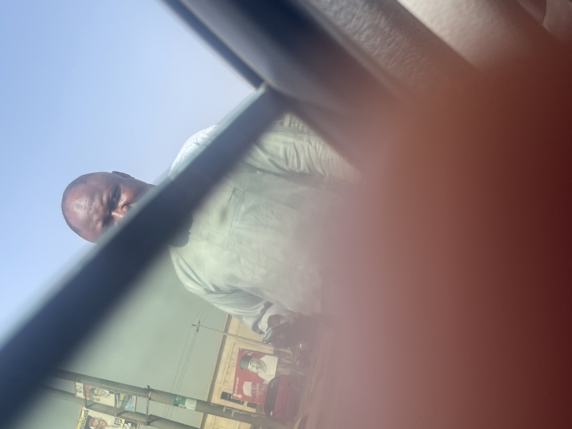 An outdoor, rotated image captures a bald man looking towards the camera, seen from inside a vehicle. The man, likely of African descent, wears a light-colored, possibly traditional, long-sleeved garment with vertical patterns. He is positioned in the lower-left quadrant of the frame against a clear blue sky, partially obscured by a dark, diagonal structure, likely a car window frame or pillar.
The lower portion of the image reveals what appears to be a street scene in bright daylight. Several political campaign posters are affixed to a pole or structure. One prominent poster features the face of a man wearing a cap, with the text "MADADA" and "20" (likely part of a year, e.g., 2023 or 2027) clearly visible. A red vehicle, possibly a small car or tricycle, is partially visible beneath these posters. Utility lines are faintly discernible against the sky in the background.
A significant portion of the right side of the image is obstructed by a large, blurry, reddish-orange object, which appears to be a finger or hand covering the camera lens. The overall impression is that of a candid shot taken from a moving or stationary vehicle in an urban or semi-urban area during the daytime, likely in Keffi, Nigeria, as provided by the context.
