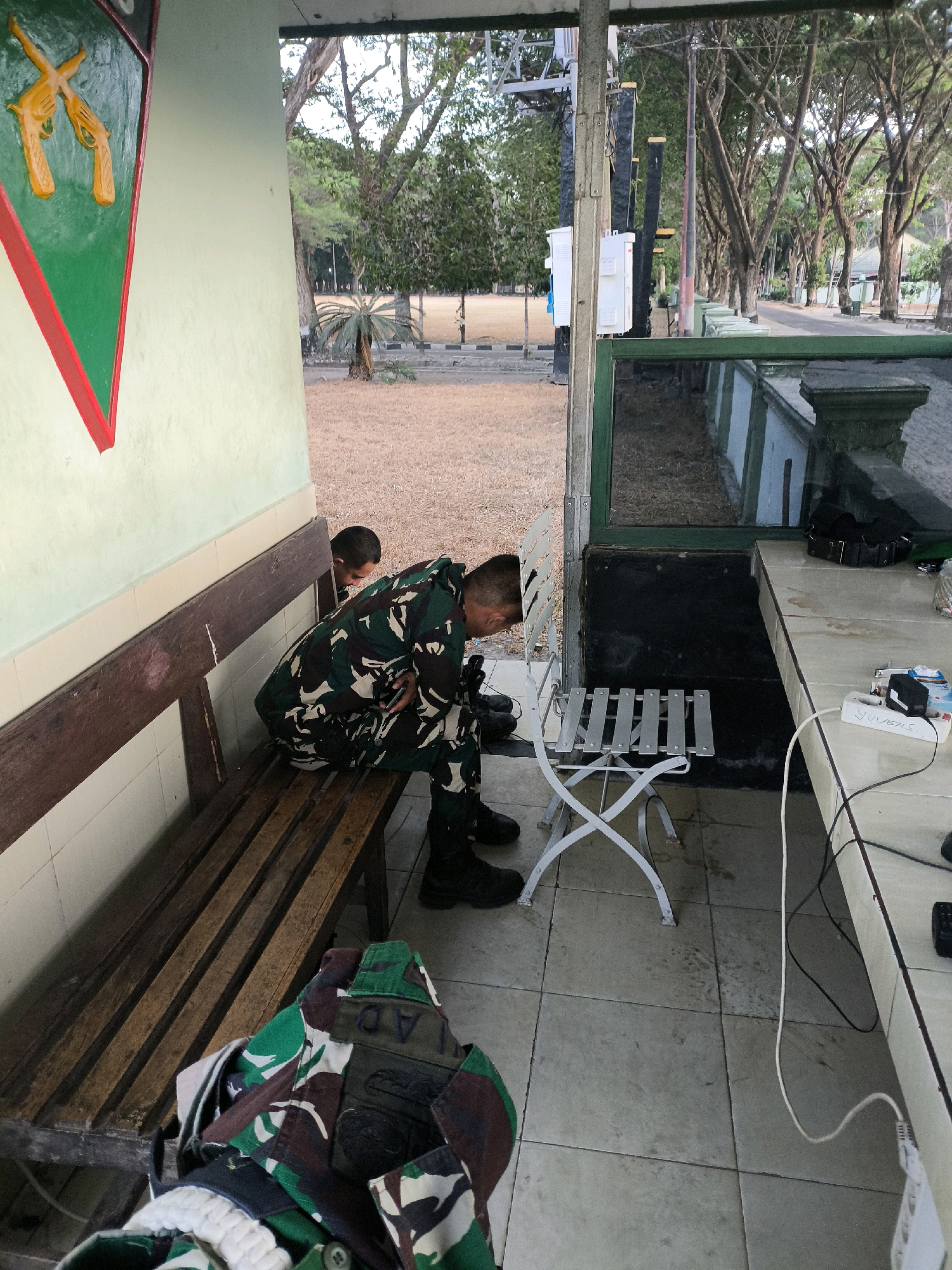 In a covered outdoor area in Unknown, Indonesia, a person in camouflage military uniform is seated on a wooden bench, looking down at their mobile phone. Another person, also in camouflage military uniform, is seated on the same bench, partially obscured by the first person, and is looking down. In the foreground, a folded camouflage military uniform jacket lies on the floor. The environment consists of a tiled floor, a wooden bench, a white metal chair, a table with electrical equipment including a power strip, and a wall with a shield emblem depicting crossed rifles. Beyond the covered area, trees and a road are visible.