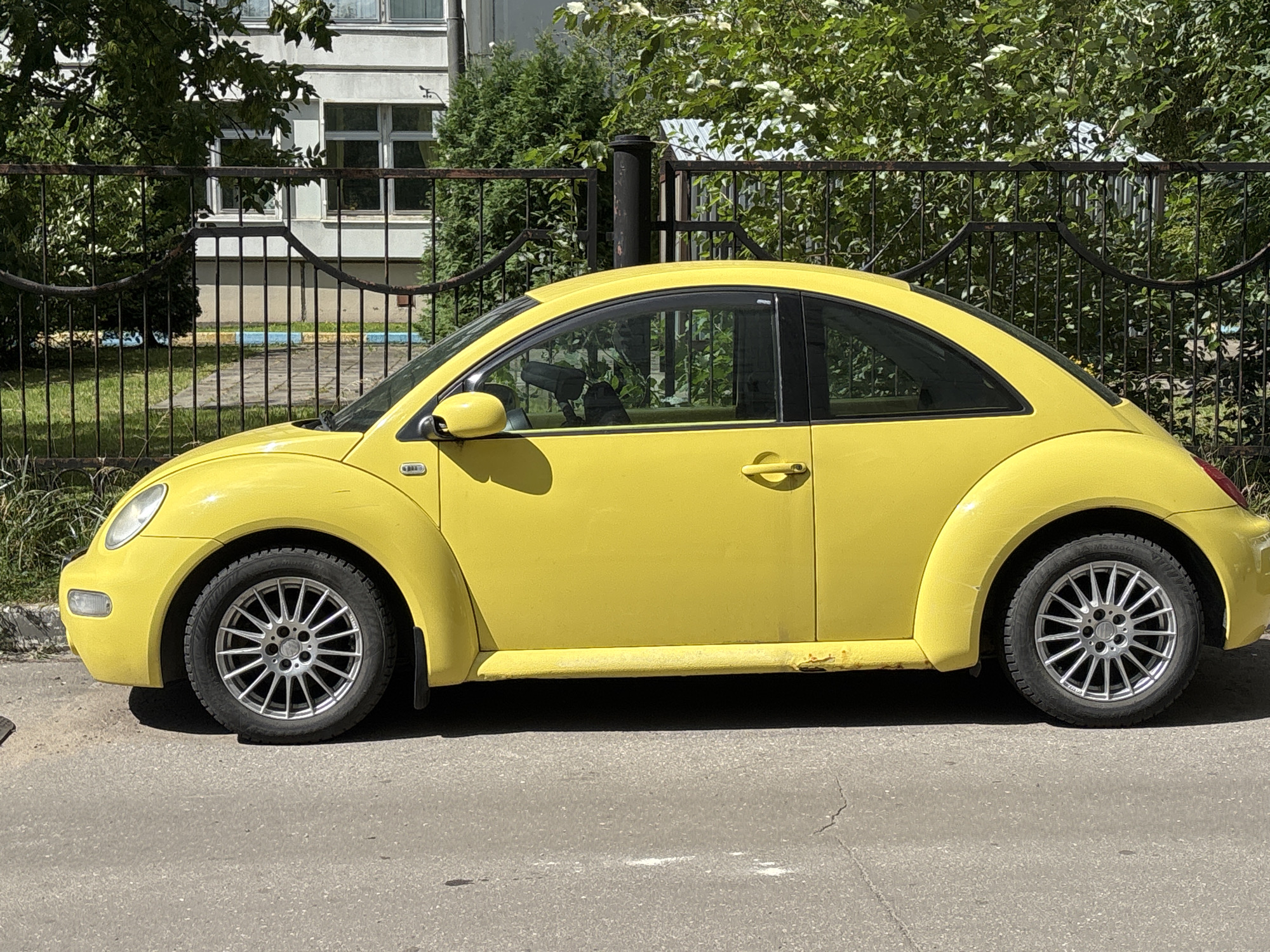 A yellow Volkswagen Beetle is parked on an asphalt road in front of a black metal fence. Behind the fence is a building with white walls and several windows, surrounded by green trees and bushes. The car is facing left, and its side is visible. The sun is shining, casting shadows on the road. The location is Odintsovo, Russia.