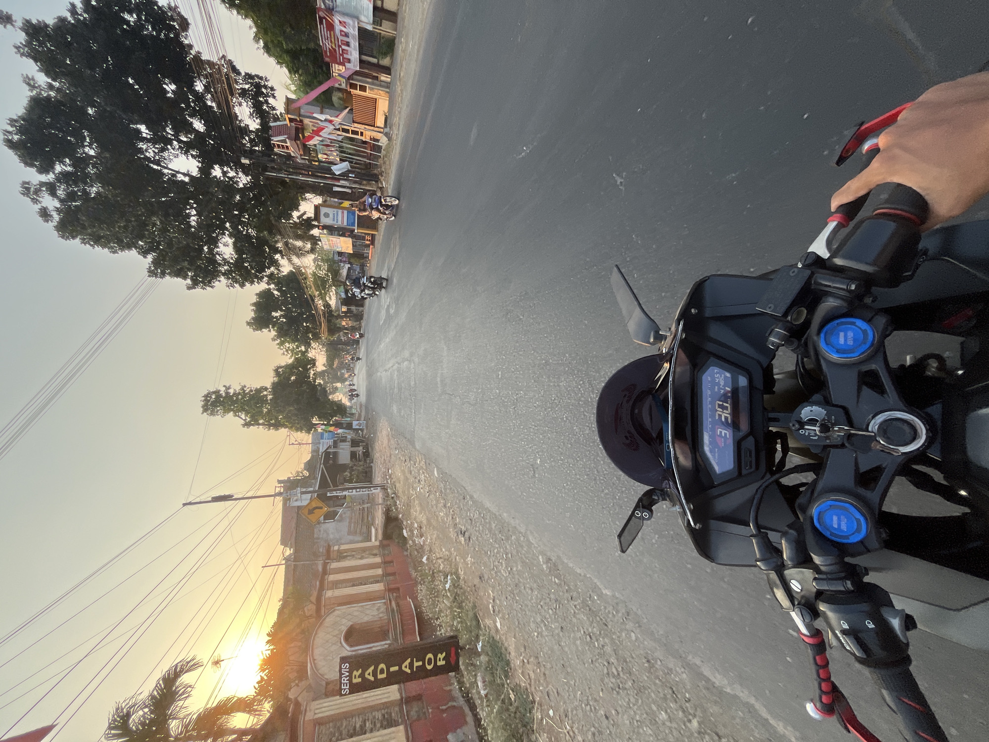 The image captures a first-person perspective of a person riding a motorcycle on a road in Sengonbugel, Indonesia, during sunset. The sun is low on the horizon, casting a warm glow over the scene. The road is paved and appears to be a local thoroughfare, with buildings and vegetation lining its sides.
In the foreground, the motorcycle's handlebars and dashboard are visible, indicating a speed of 30 km/h. The dashboard display shows the time as 4:57. The rider's left hand is on the grip, and their right hand is visible reaching for the throttle.
Along the left side of the road, a red and white building with arched windows stands, and a vertical sign reads "SERVIS RADIATOR" in black lettering. Palm trees and other foliage are present.
On the right side of the road, there are more buildings, some adorned with Indonesian flags, suggesting a local celebration or event. A large tree with dense foliage is prominent. Several other motorcycles and riders are visible further down the road, moving in the same direction as the viewer. The sky is a pale orange and yellow, characteristic of a sunset. The overall impression is one of a peaceful commute through a rural or semi-urban Indonesian town.
