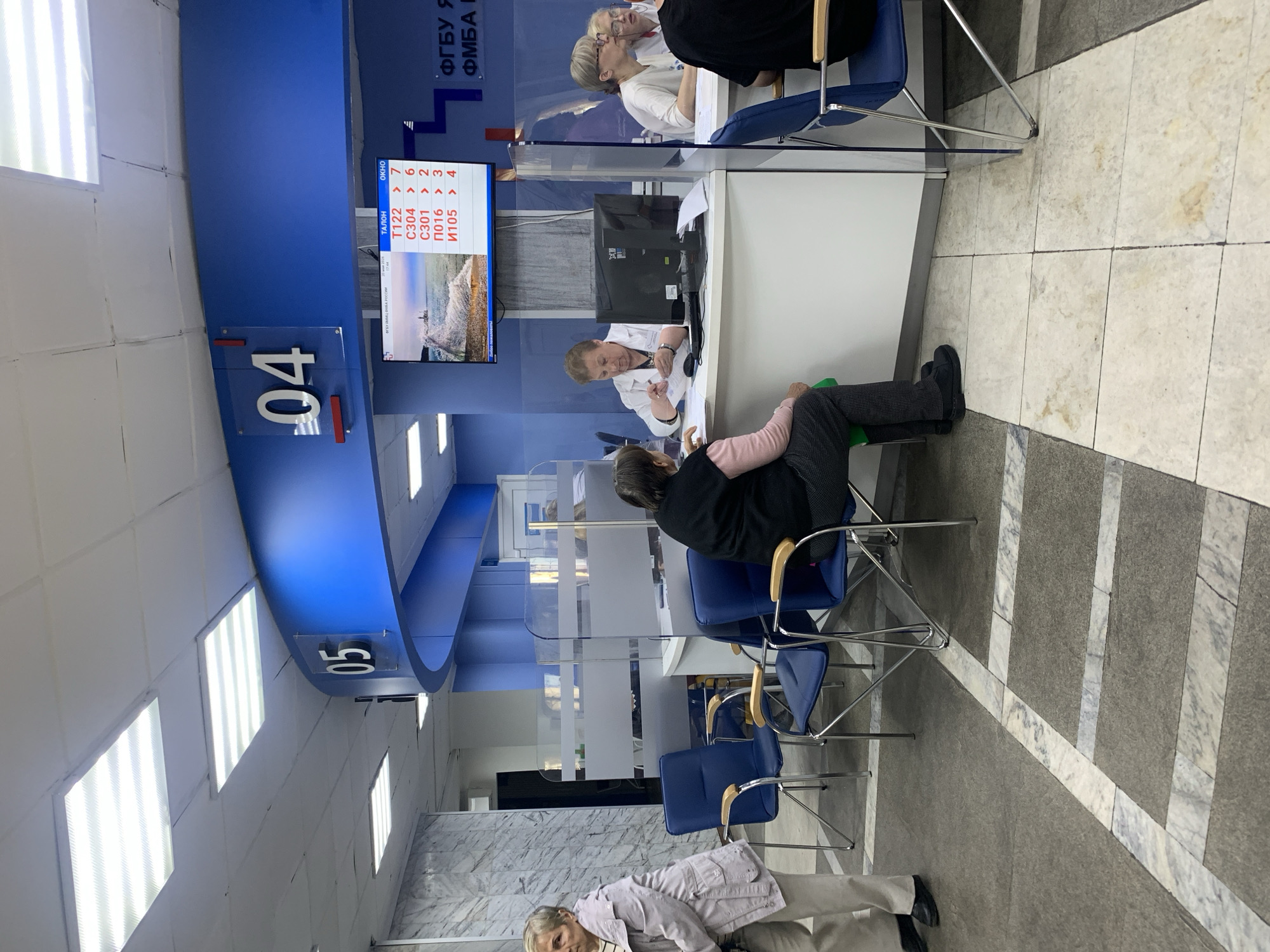 The interior shot depicts a waiting area inside an office, presumably in Unknown, Unknown. A dark grey and white marble-patterned floor dominates the bottom portion of the frame.
Along the left edge, blue numbers "05" and "04" are affixed to the ceiling, indicating the service counters. A blue band spans the upper portion of the image, and above this band a display screen shows the next numbers to be served: T122, C304, C301, П016 and И105.
Several people are present. Two women are seated behind counters, each with a computer monitor. Clear plexiglass partitions separate them from the customers. Two seated customers are visible: one is actively being served, and the other is waiting.
Chairs with blue seats are arranged in rows for waiting customers. The scene appears to be within a public service office, perhaps a government agency or a healthcare facility. The room is illuminated by fluorescent lights on the ceiling.