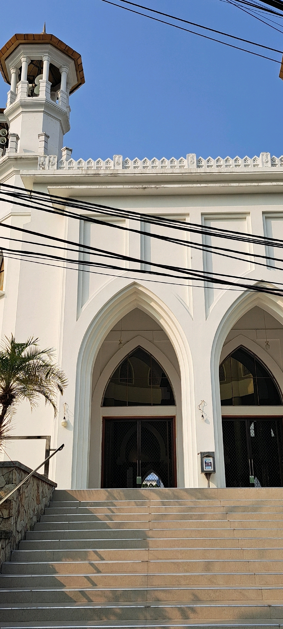 The image shows the entrance to a white building, likely a mosque, in the city of Petungasri, Indonesia. A wide set of stone stairs leads up to two large arched doorways, each with dark double doors. Above the doorways are arched windows with reflective glass. The building has decorative white trim along the top. A minaret, also white with a copper-colored roof, is visible on the left side of the building, with what appear to be loudspeakers mounted on it. A palm tree stands to the left of the stairs. Electrical wires crisscross the upper portion of the image against a clear blue sky. The scene is bathed in sunlight, casting shadows on the stairs. There are no people visible in the image.