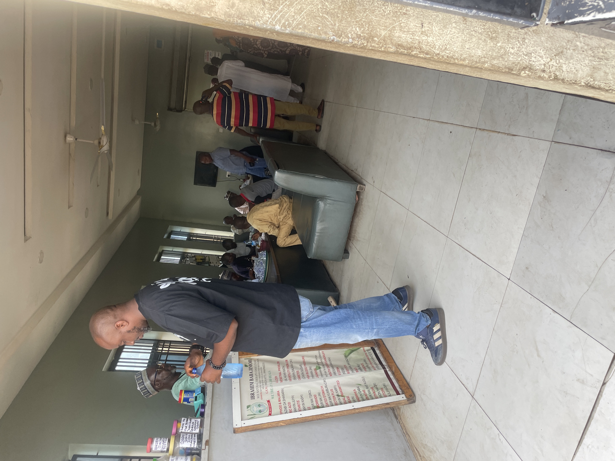 The image depicts an indoor scene that appears to be a waiting area or reception of a natural therapy clinic in Bauchi, Nigeria. In the foreground, a bald man wearing a black t-shirt and blue jeans is holding a plastic bottle of water. He is standing next to a wooden board with a list of ailments and treatments written on it, titled "IBRAHIM BABA JIMOH, N.D. NATURAL THERAPY CONSULTANT." The board is positioned on an easel.
In the background, several other individuals are visible. To the left, a man wearing glasses and a cap is behind a counter, interacting with the man in the foreground. Further back, there are people sitting on a green sofa and standing. One man in a striped shirt is on his phone. The room has tiled floors and a white ceiling with ceiling fans. A television is mounted on the wall. The overall environment suggests a functional, albeit somewhat dated, establishment. The natural light filtering in from the left suggests the entrance or a window is in that direction.
