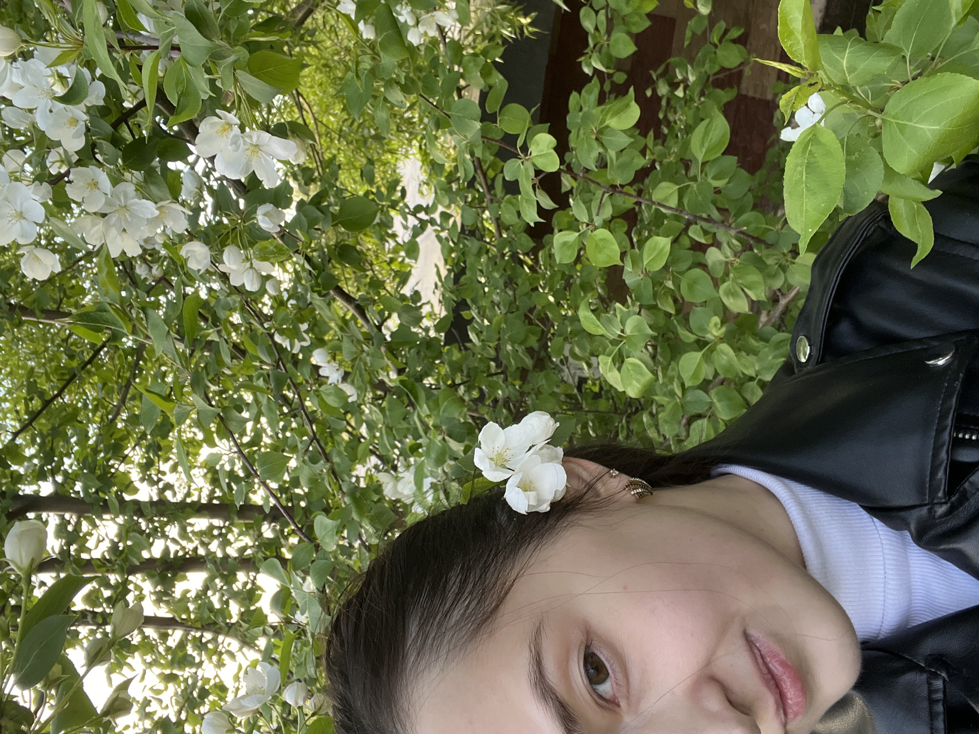 A close-up shot shows a woman from the nose up. She has brown hair and is wearing an earring. A white flower sits above her ear. She is wearing a white undershirt and a black leather jacket. The background is a variety of green leaves, with some white flowers in the background as well. The location appears to be outdoors. There is no indication this image was taken in the city of Dzerzhinsk, Russia.