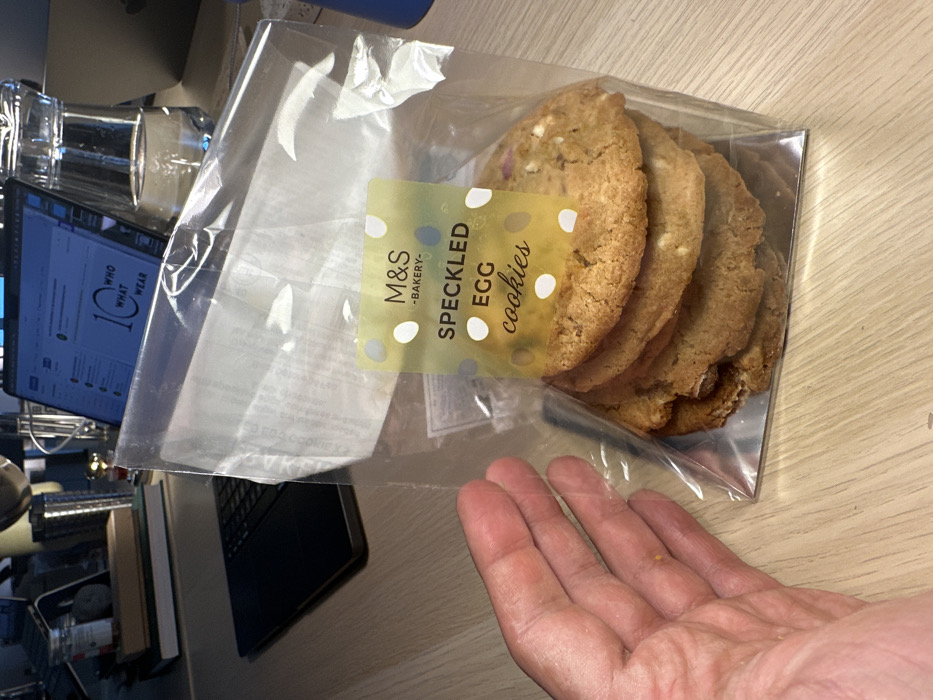 The scene depicts an indoor office or home office environment in London, United Kingdom.
A clear plastic bag containing a stack of approximately five to six "M&S BAKERY SPECKLED EGG cookies" is the central subject, resting on a light-colored wooden desk surface. The cookies are round, light brown, and contain visible white inclusions. The bag features a yellow-green label with white and light green polka dots, bearing the text "M&S -BAKERY- SPECKLED EGG cookies". A silver-colored cardboard insert forms the base of the cookie packaging.
To the left of the cookie package, a bare human left hand, with fingers slightly splayed, rests horizontally on the desk, appearing to hold or interact with the bag.
The background reveals additional objects on the desk. A dark-colored laptop with a black keyboard is open to the left, displaying a website with "10 WHO WHAT WEAR" visible on its screen. Behind the laptop, a small stack of books or notebooks, a clear glass jar with a red lid, a metallic cylindrical object, and a small, dark, irregularly shaped object are arranged. To the right of the laptop and behind the cookie package, a clear glass carafe or jug filled with water is present. A blue plastic bottle or cup is partially visible on the far right of the frame. The lighting suggests an artificial source, typical of an interior workspace.