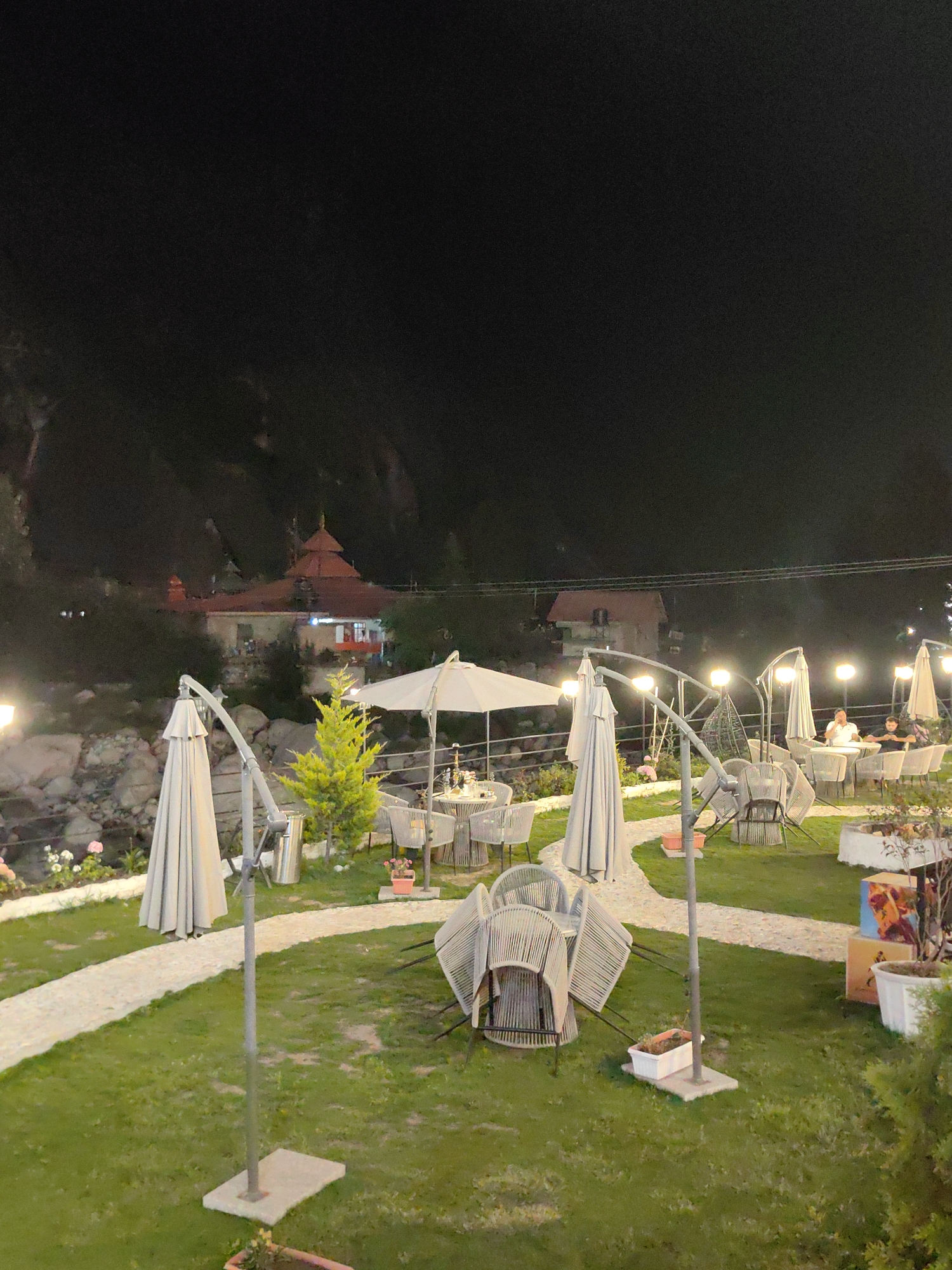 The image depicts an outdoor seating area at night, likely a restaurant or cafe, situated near a mountainous landscape and possibly a body of water. In the background, a building with a red, tiered roof, resembling a temple or traditional Indian architecture, is visible. The foreground is dominated by a well-maintained lawn with several white patio umbrellas and a curved, light-colored pathway. Outdoor furniture, including chairs and tables, is arranged on the lawn and pathway. Some chairs are stacked. Several lampposts with bright lights illuminate the area. In the mid-ground, a small, bright green evergreen tree stands near a cluster of tables. Two individuals are seated at one of the tables in the mid-ground, appearing to be engaged in conversation. The overall impression is a serene and inviting atmosphere for evening dining or relaxation in Kulang, India.