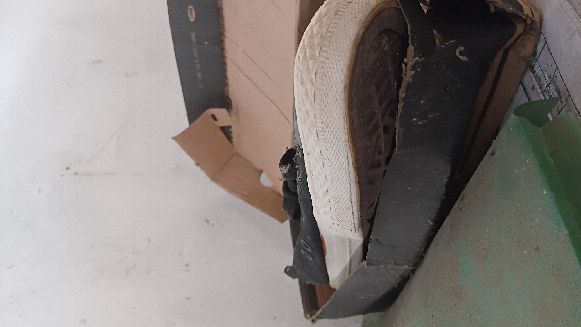 A black shoe with a white sole sits in a damaged black and brown cardboard box. The box appears to be old and worn, with rips and tears in the material. The shoe is partially visible, with the toe and side facing upwards. The background is a plain, white wall. This scene could be found in a storage area or a neglected corner of a home in Prabumulih, Indonesia.