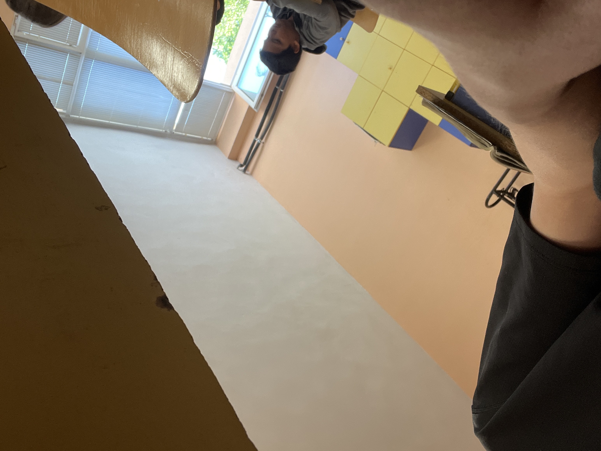The image is taken from a low angle, looking upwards in a room that appears to be a school or educational facility. A person wearing a gray shirt is visible looking down in the distance. A table is in the foreground, and the room contains a window with blinds. The walls are peach colored. There are yellow square objects. A person with a black shirt appears to be taking the picture. This location is probably not in the city of Kavarna, Bulgaria.