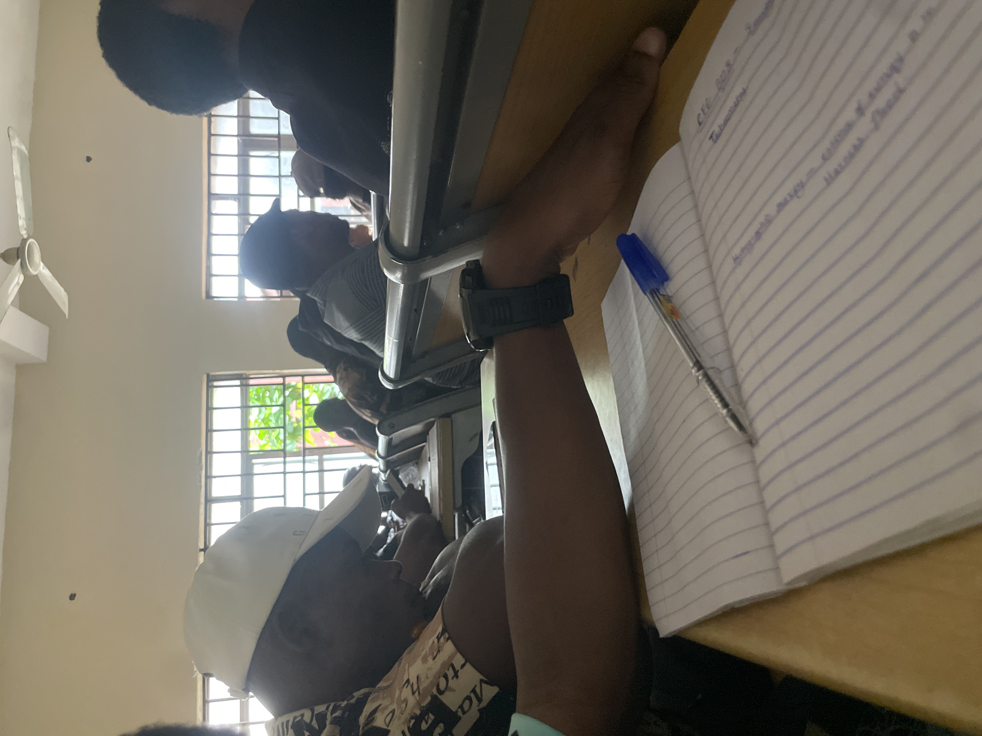 The image depicts a classroom setting, likely within the city of Ihiagwa, Nigeria, given the context. In the foreground, a notebook with lined pages is open on a wooden desk. A blue ballpoint pen rests on the notebook, with its tip pointing towards the left page. A person's right arm, adorned with a dark digital watch on the wrist, is extended across the notebook, with their hand resting on the desk's edge. This individual is wearing a patterned shirt.
In the background, other individuals are seated at desks, some wearing hats and casual attire. The classroom is illuminated by natural light filtering through windows with barred frames, suggesting a secure environment. A ceiling fan is visible on the upper portion of the frame. The overall scene conveys a typical academic atmosphere, potentially during a lecture or study session. The specific location within Ihiagwa cannot be precisely determined from the image alone, but the visual cues align with educational institutions found in the region.