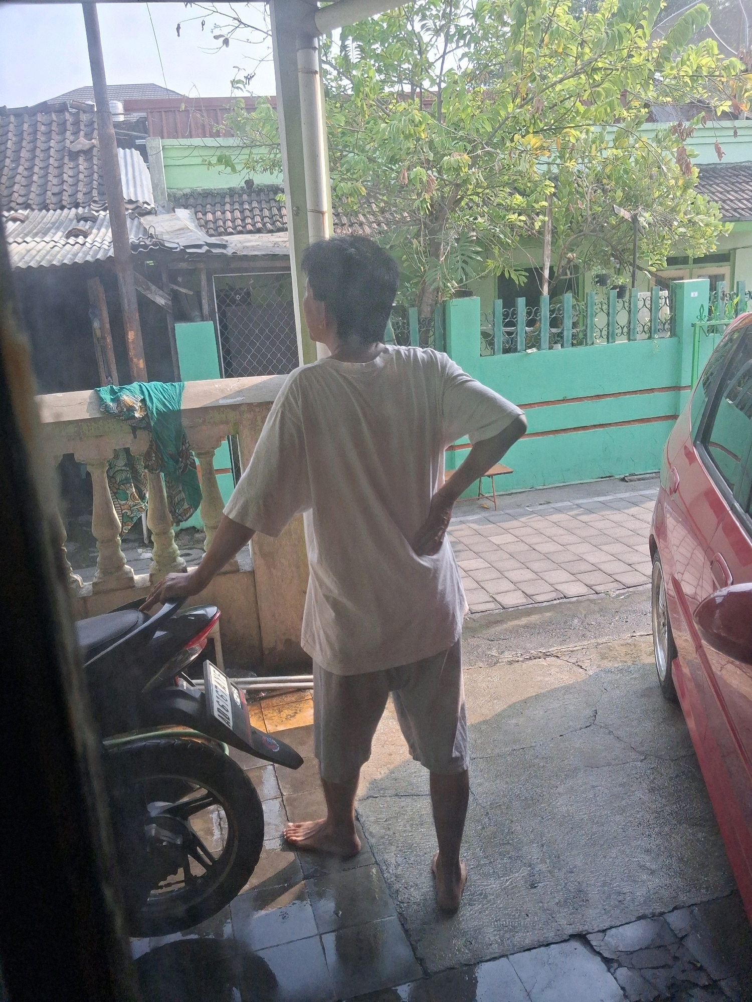 A person stands facing away from the camera, leaning against a motorcycle. They are wearing a white t-shirt and light-colored shorts. The motorcycle is dark colored and parked on a tiled surface that appears wet, possibly from recent rain. To the right of the motorcycle, a portion of a red car is visible. In the background, there is a green fence, trees, and buildings with tiled roofs, consistent with the architecture found in Surakarta, Indonesia. The overall scene suggests an outdoor residential or alleyway setting.