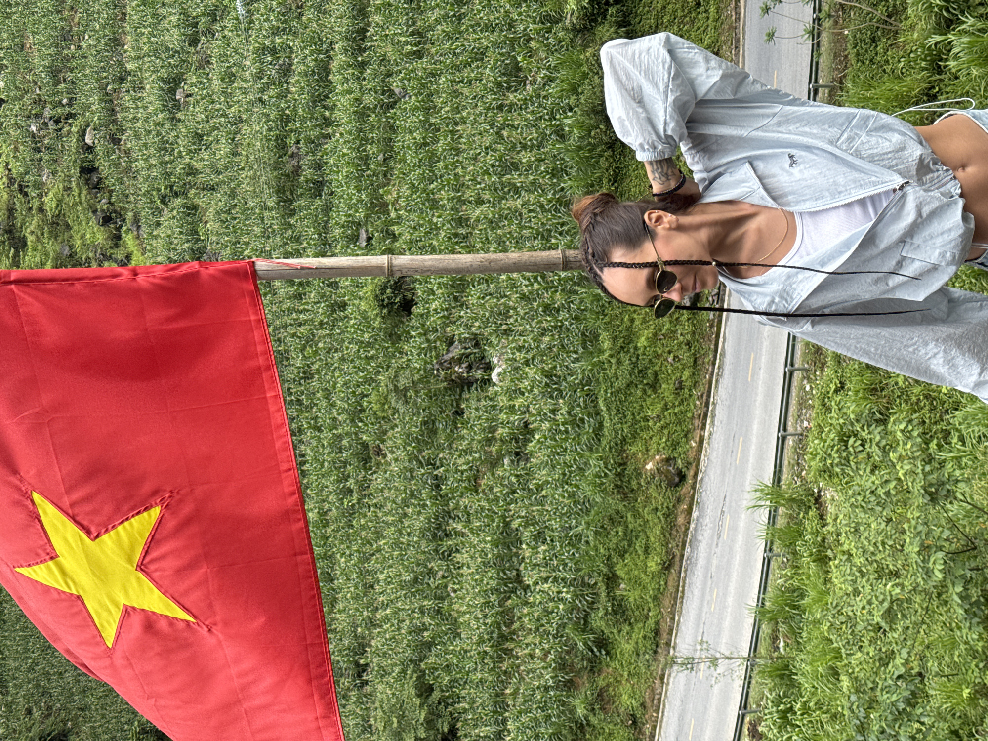 A vertical, medium shot captures a woman posing beside a Vietnamese flag against a backdrop of lush greenery in Yên Minh Commune, Vietnam. The flag, predominantly red with a central yellow star, is positioned on the left side of the frame. The woman stands on the right, wearing sunglasses, a white top, and a light blue jacket. A paved road runs along the edge of the greenery behind her. The dense vegetation, likely part of the landscape of Yên Minh, fills the majority of the background.