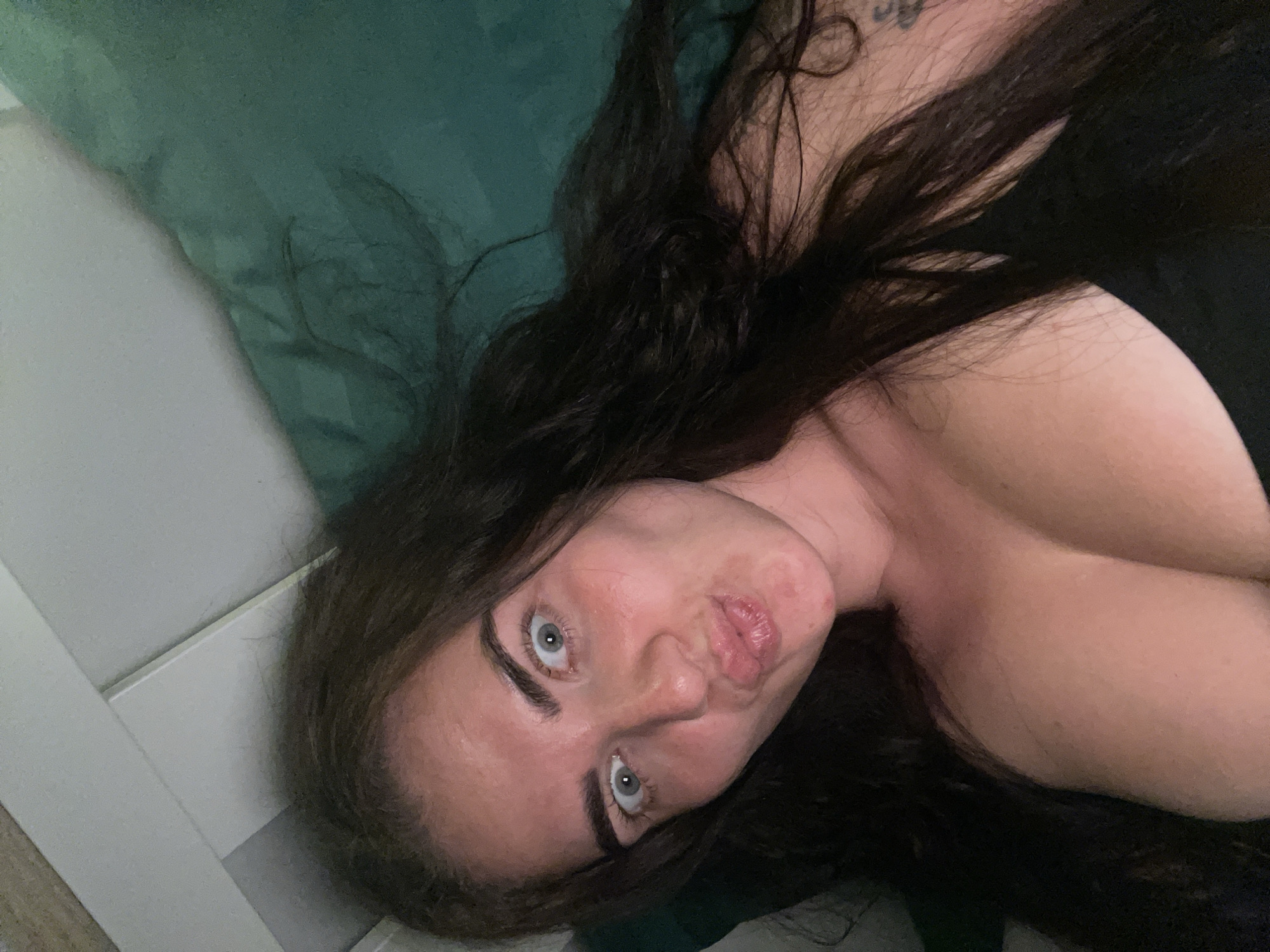 The image shows a close-up, high-angle shot of a woman lying down. She has long, dark brown hair and is looking directly at the camera with wide, light blue eyes. Her lips are puckered as if for a kiss, and her eyebrows are notably dark and well-defined. She appears to be wearing a black top, with her chest partially visible. The background consists of a green pillow and a white surface, possibly a headboard or wall. The lighting suggests an indoor environment, likely a bedroom. There is no indication of the location being in Мосметаллоконструкция Онт, Russia.