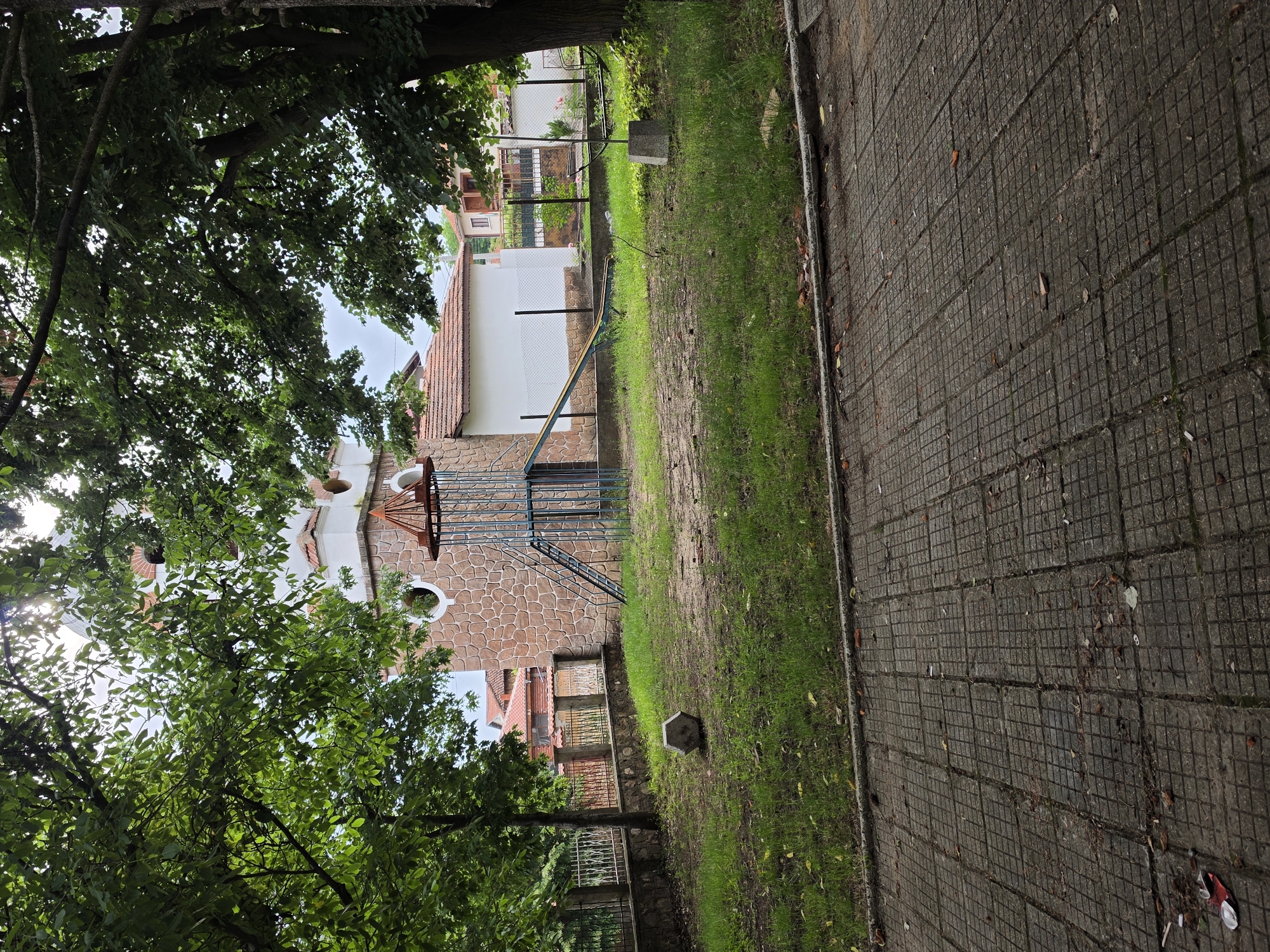 The image depicts a children's playground area in Turiya, Bulgaria, situated in front of a stone-clad building with a prominent tower. The playground features a blue and yellow slide and a climbing structure. The ground is covered with patchy grass and dirt. A concrete pathway, composed of square tiles, runs along the foreground. Surrounding the playground is a fence, and behind it, residential buildings with red-tiled roofs are visible. Mature trees with lush green foliage arch over the scene, partially obscuring the sky. A small, hexagonal stone object rests on the grass in the middle ground. The overall impression is one of an established, perhaps somewhat aged, community space.