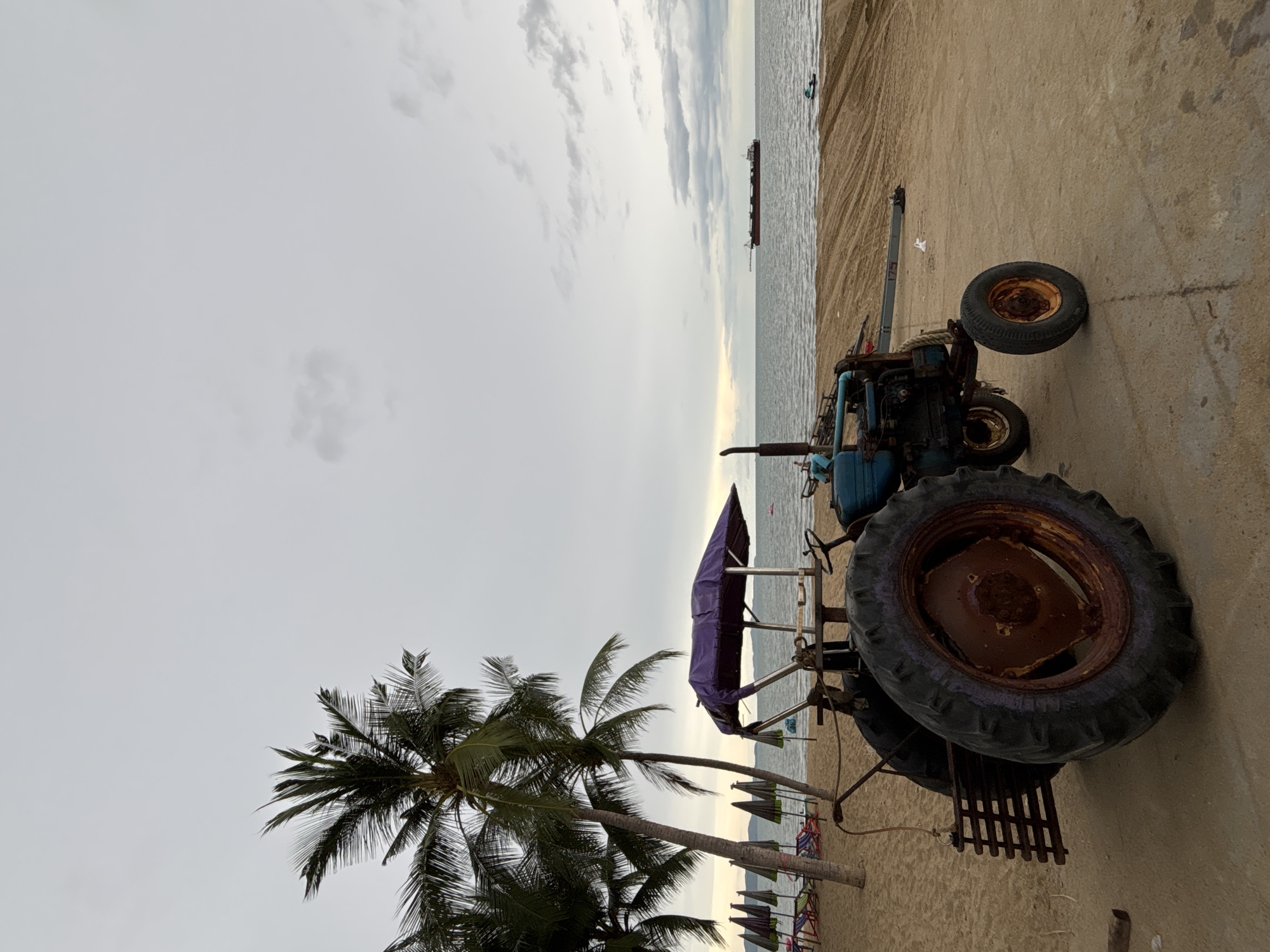 This is a full-frame, eye-level, vertical shot of a beach scene in Pattaya City, Thailand, under a cloudy sky. In the foreground, a weathered blue and rust-colored tractor is parked on the sand and concrete. The tractor has large, worn rear tires with purple accents and a smaller front tire. A purple canopy is attached to the tractor. Behind the tractor, a sandy beach stretches towards the ocean. Several beach umbrellas and lounge chairs are visible to the left, lined up near palm trees. In the distance, a large cargo ship is sailing on the calm sea. The overall atmosphere is tranquil and slightly overcast.