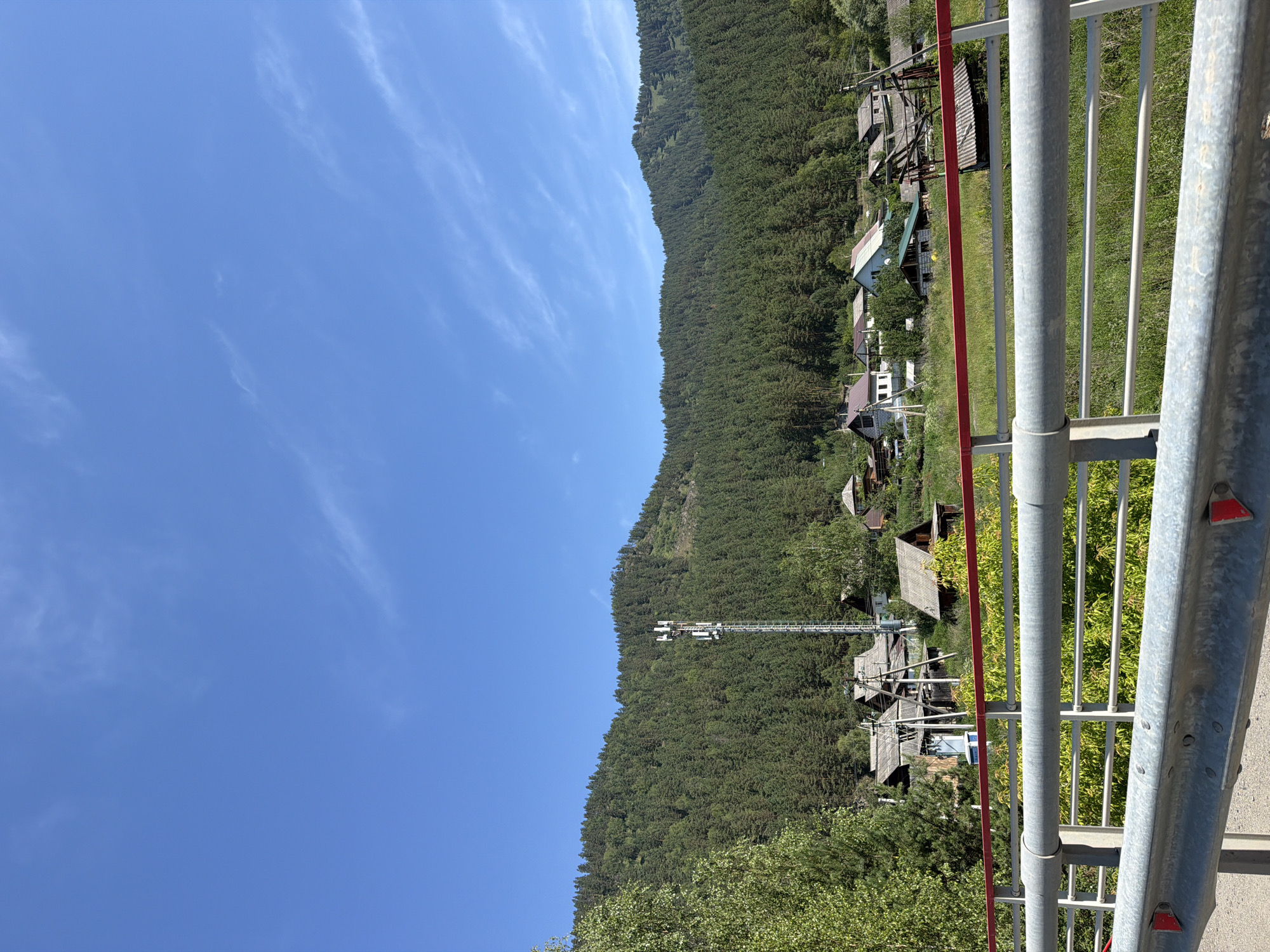 The image shows a view from a bridge overlooking a village nestled at the base of a heavily forested mountain. The sky is a clear, bright blue with a few wispy white clouds. The village consists of several houses with wooden roofs, some appearing to be older or in disrepair, scattered amongst trees and vegetation. A tall cell tower stands prominently in the middle ground, equipped with multiple antennas. The foreground features the metal railings of the bridge, with red and white reflective markers visible on the lower barrier. The surrounding environment is lush and green, with dense coniferous trees covering the mountainside and lighter foliage on the lower slopes. The overall impression is of a rural, somewhat remote settlement in a natural, mountainous setting. The scene is consistent with the location of Ust'-Muny, Russia, known for its mountainous terrain and natural beauty.
