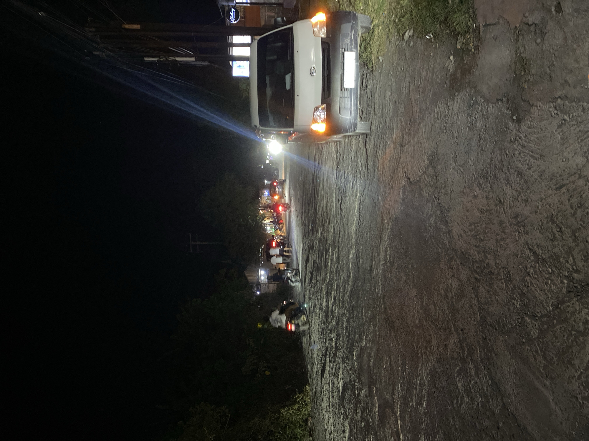 The image depicts a nighttime scene on a paved road in Kuta Selatan, Indonesia. A white van is parked on the right side of the road, its headlights illuminated. Several motorcycles are visible on the road, some with riders, suggesting movement or a gathering. In the background, there are trees and some illuminated buildings or stalls. The road surface appears somewhat rough and uneven. Power lines and poles are present on the right side of the image. The overall atmosphere is that of a busy street at night.