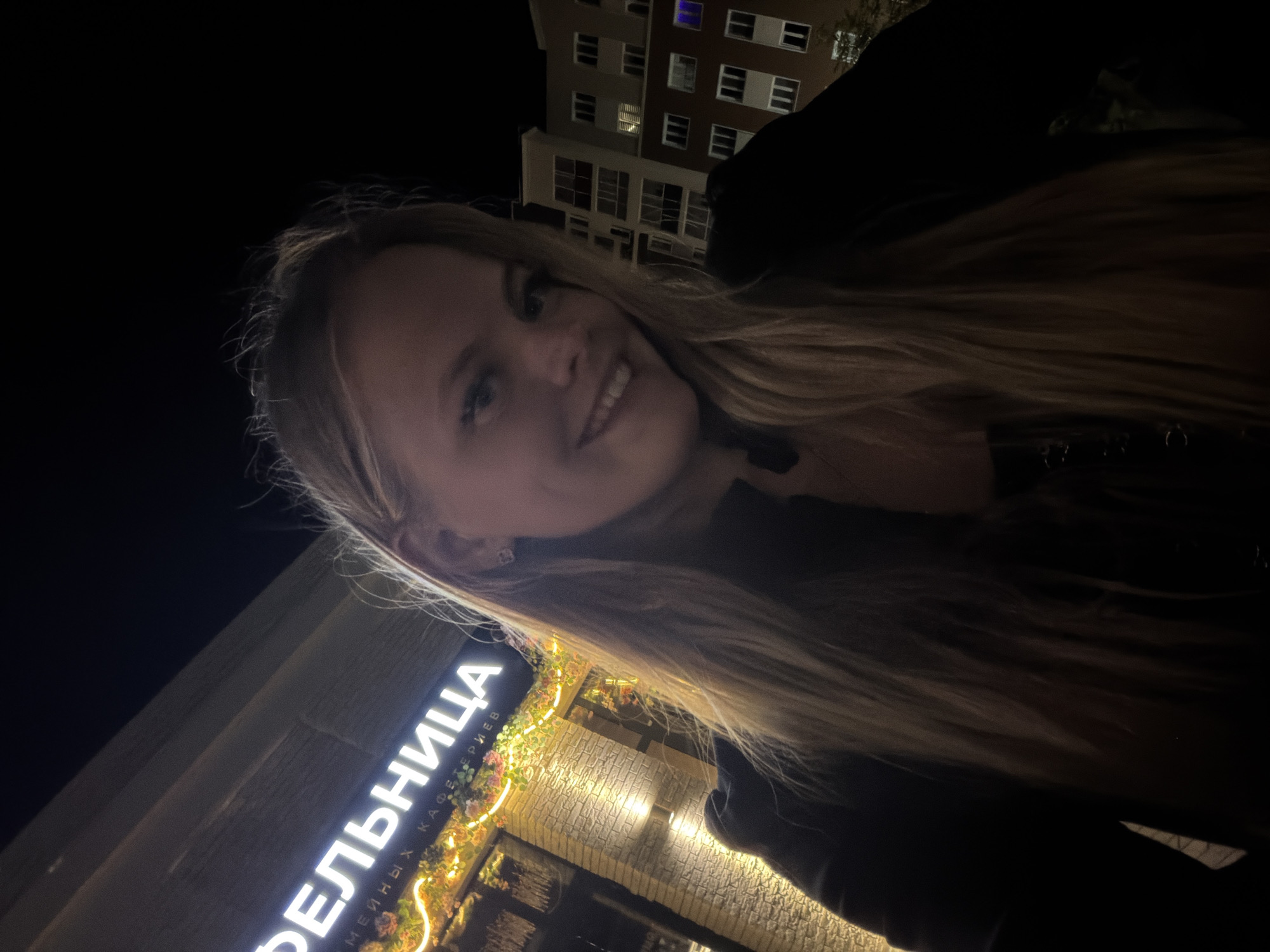 A young woman with long blonde hair and a black top smiles at the camera. She is standing outside a building at night. The building has a sign that reads "БЕЛЬНИЦА" in large white letters, and is decorated with string lights and artificial flowers. Behind her, an apartment building is visible. The image appears to have been taken in Pervomaysky, Russia.