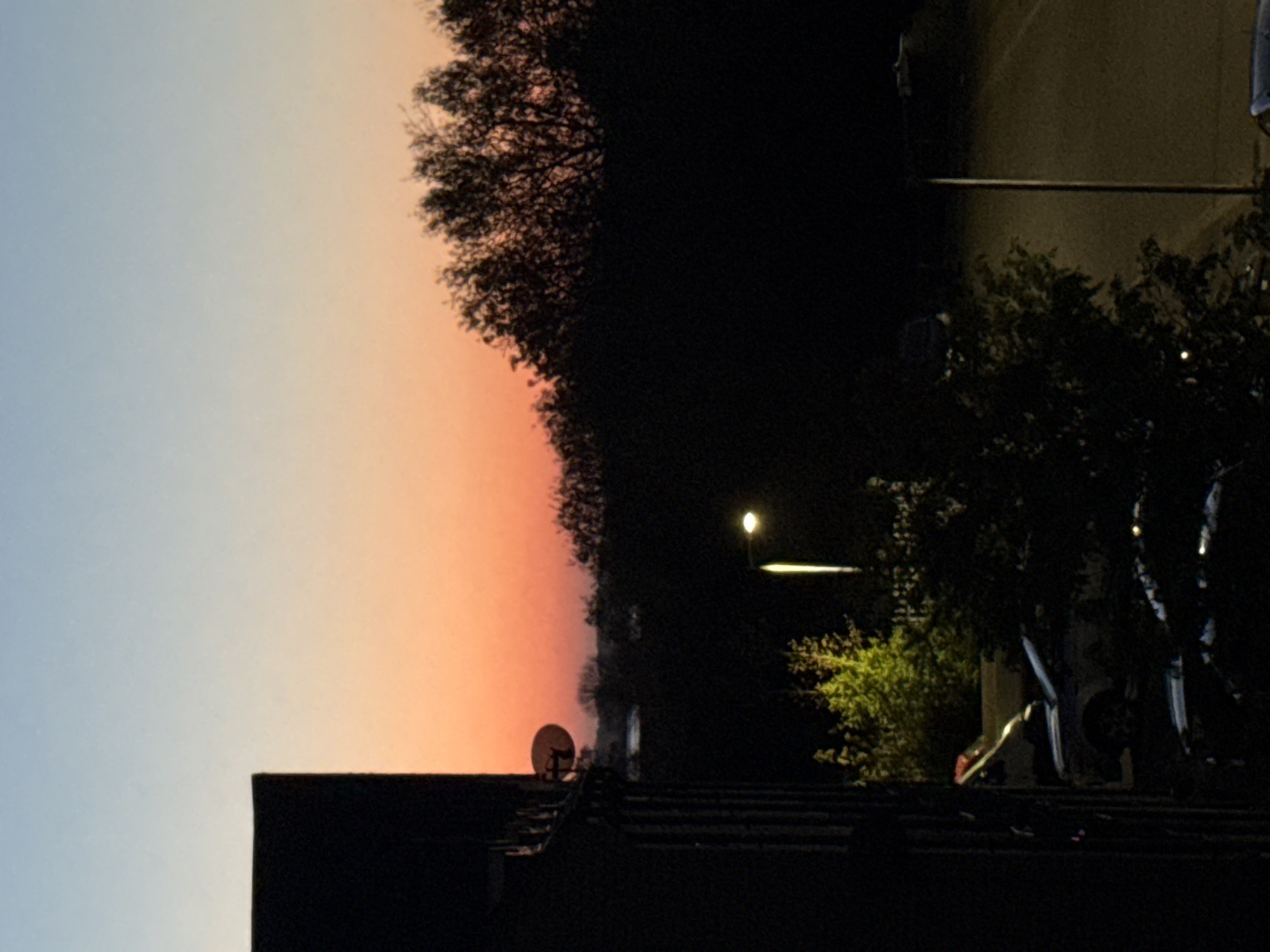 The image depicts a twilight scene in Great Yarmouth, United Kingdom. The sky transitions from blue to orange and pink hues. A dark tree line obscures much of the upper-right portion of the image. Below the skyline, the dark roof of a building is visible, with a satellite dish mounted on top. A street lamp illuminates a road and adjacent trees. Parked bicycles are visible on the right side of the frame. The overall lighting conditions are low.