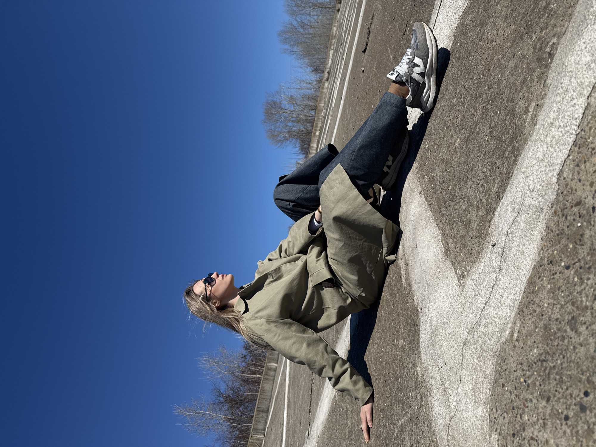 A young woman with fair hair is lying on a concrete surface under a clear blue sky. She wears sunglasses, a light olive-green trench coat, and dark blue pants. A pair of grey and white New Balance sneakers are on her feet. The concrete surface has painted white lines. Bare trees and vegetation are visible in the background. The image was taken on a clear day in Chaikovsky, Russia.