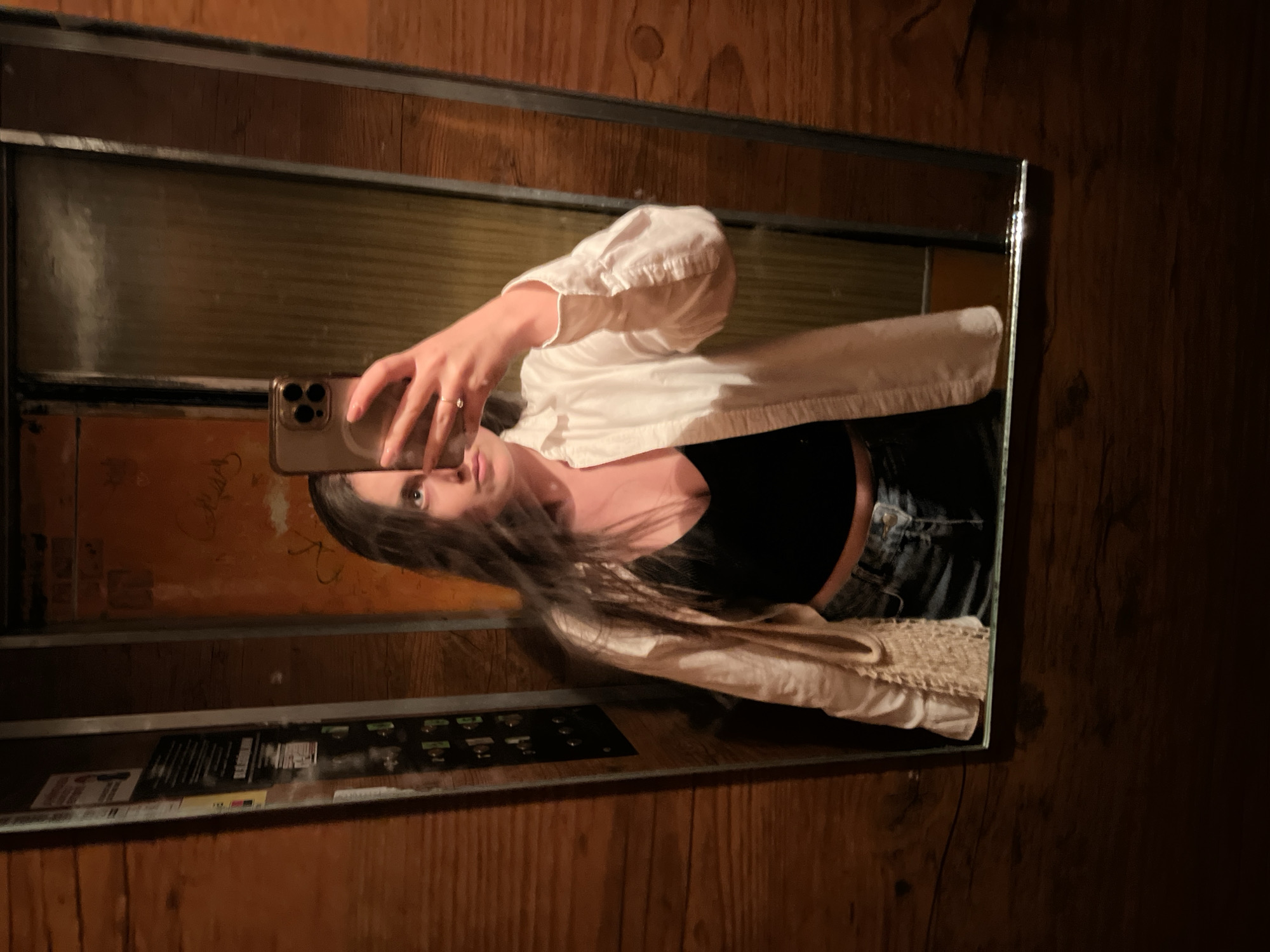 A person is taking a mirror selfie in what appears to be an elevator. The person has long, dark hair and is wearing a black top, jeans, and a white shirt or jacket. They are holding a smartphone in front of their face to take the picture. The background shows the elevator's interior, including buttons, paneling, and other informational displays. The elevator is located in Ternopil, Ukraine. The walls outside the elevator appear to be wooden.