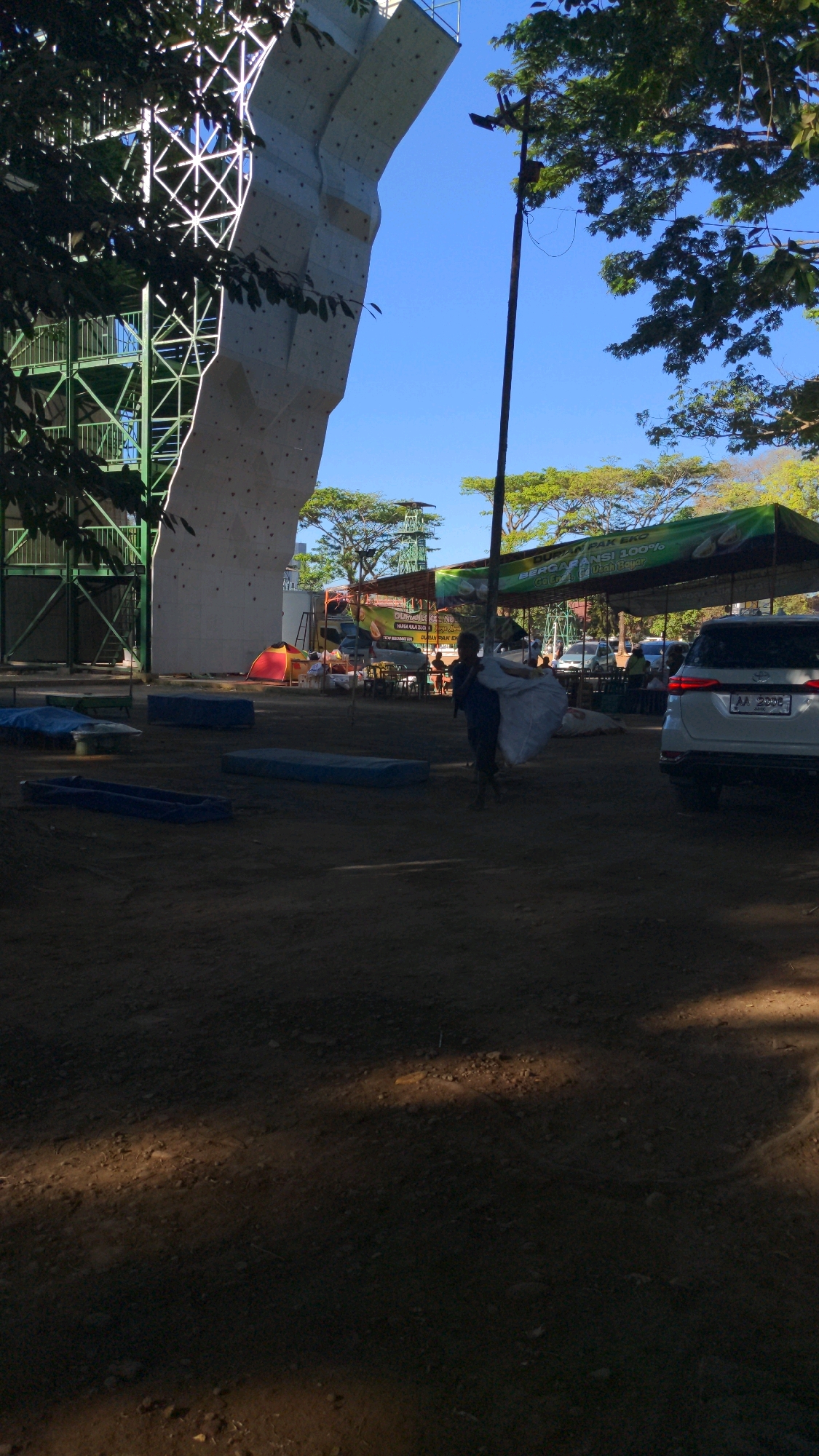 A clear, sunny day in Magelang, Indonesia, showcases an outdoor climbing wall with a complex metal support structure. In the foreground, scattered mats and tarps lie on a dirt ground. A young person, silhouetted against the bright light, carries a large bundle of white material. Behind them, a white SUV is parked, and further in the background, tents and stalls are visible under a green canopy, suggesting a marketplace or event. Trees line the horizon under a vibrant blue sky.