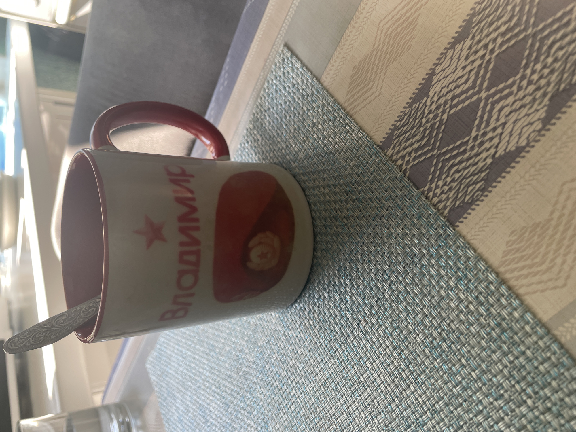 A close-up shot shows a white ceramic mug with a red rim and handle, resting on a blue and white woven placemat on a table. The mug features red Cyrillic lettering that reads "Владимир" (Vladimir) and a red emblem with a golden star and hammer and sickle. A decorative silver spoon is placed inside the mug. In the background, a glass of water and a blurred seating area are visible. The image is taken in natural light, possibly from a window, casting soft shadows. The scene appears to be indoors, in a dining or living area. No specific location in Chaikovsky, Russia, can be identified from the visual information alone.