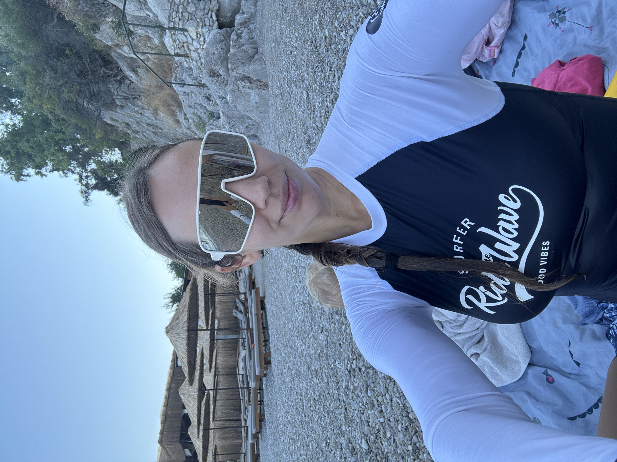 A woman is taking a selfie on a pebble beach in Fethiye, Turkey. She is wearing a black and white rash guard and large white sunglasses. Behind her, there are rows of empty lounge chairs and straw umbrellas, and a rocky hillside with some trees. A toddler with blond hair is partially visible behind her. The sky is clear and blue.