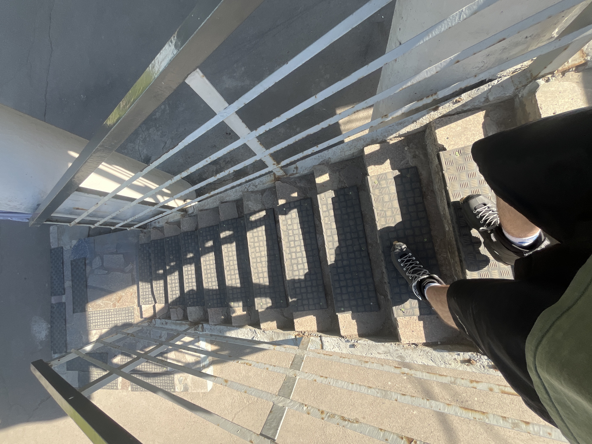 The image presents a high-angle view looking down a flight of outdoor stairs, with a person standing at the top right. The visible parts of the person include their legs, wearing black shorts, and black sneakers with white laces and white socks. A hint of a green garment is visible on their upper right.
The stairs are made of concrete, with black, textured anti-slip mats affixed to each tread. Some mats feature a diamond plate pattern, while others have a grid texture. A metal railing, painted a light grey or white, with horizontal bars, runs down both sides of the staircase. Some sections of the railing show signs of rust.
Bright sunlight casts strong, distinct shadows of the railing across the stairs and the ground below, indicating clear, sunny daytime weather, likely midday or early afternoon. The ground at the bottom appears to be a mix of concrete, asphalt, and possibly stone pavers. A light-colored wall or surface is visible on the far left. No discernible emotions are conveyed, and no text is visible in the image. The provided context for the location is Krasnoyarsk, Russia.